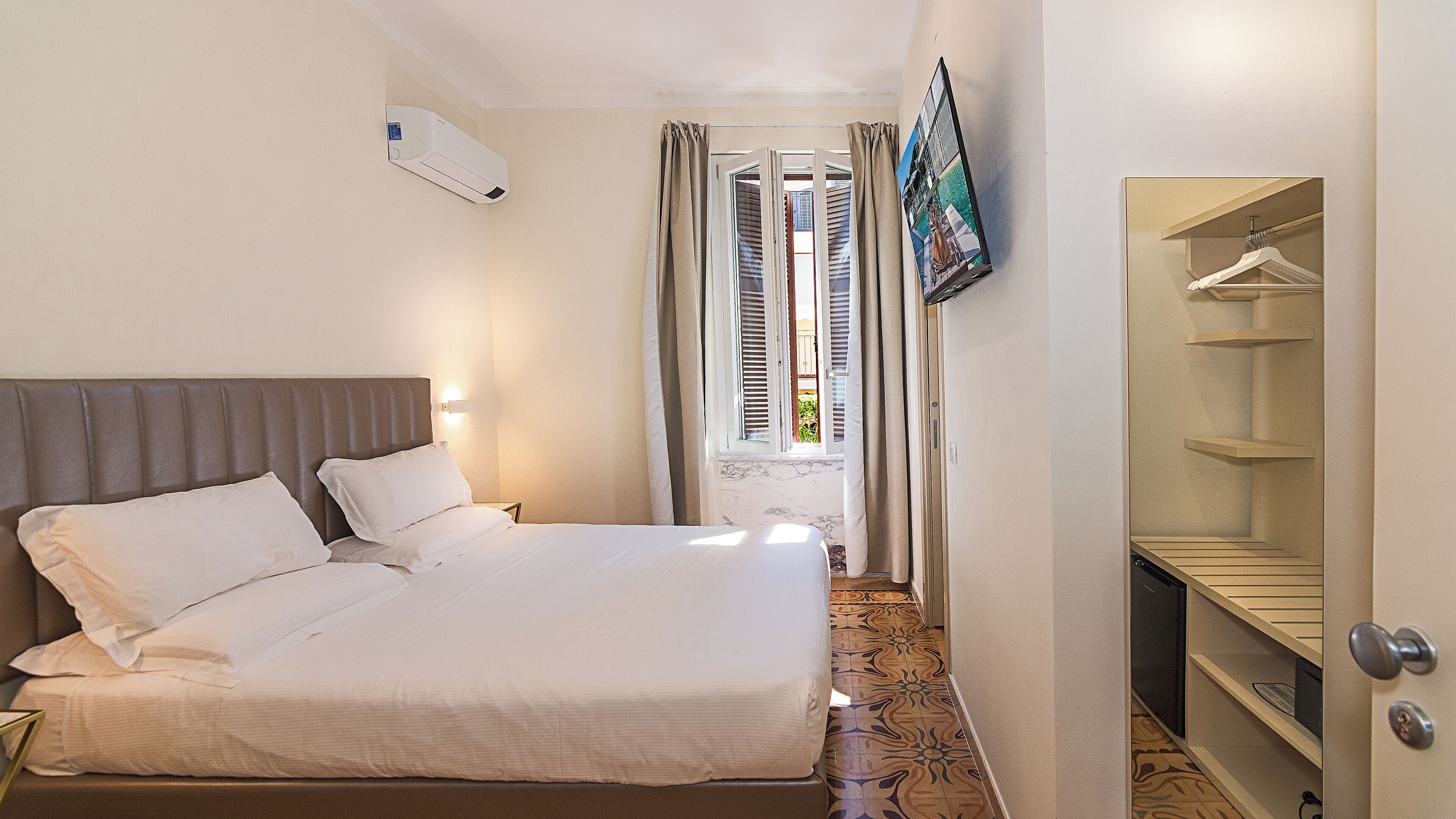Una habitación de hotel con cama, armario, ventana con cortinas y unidad de aire acondicionado.