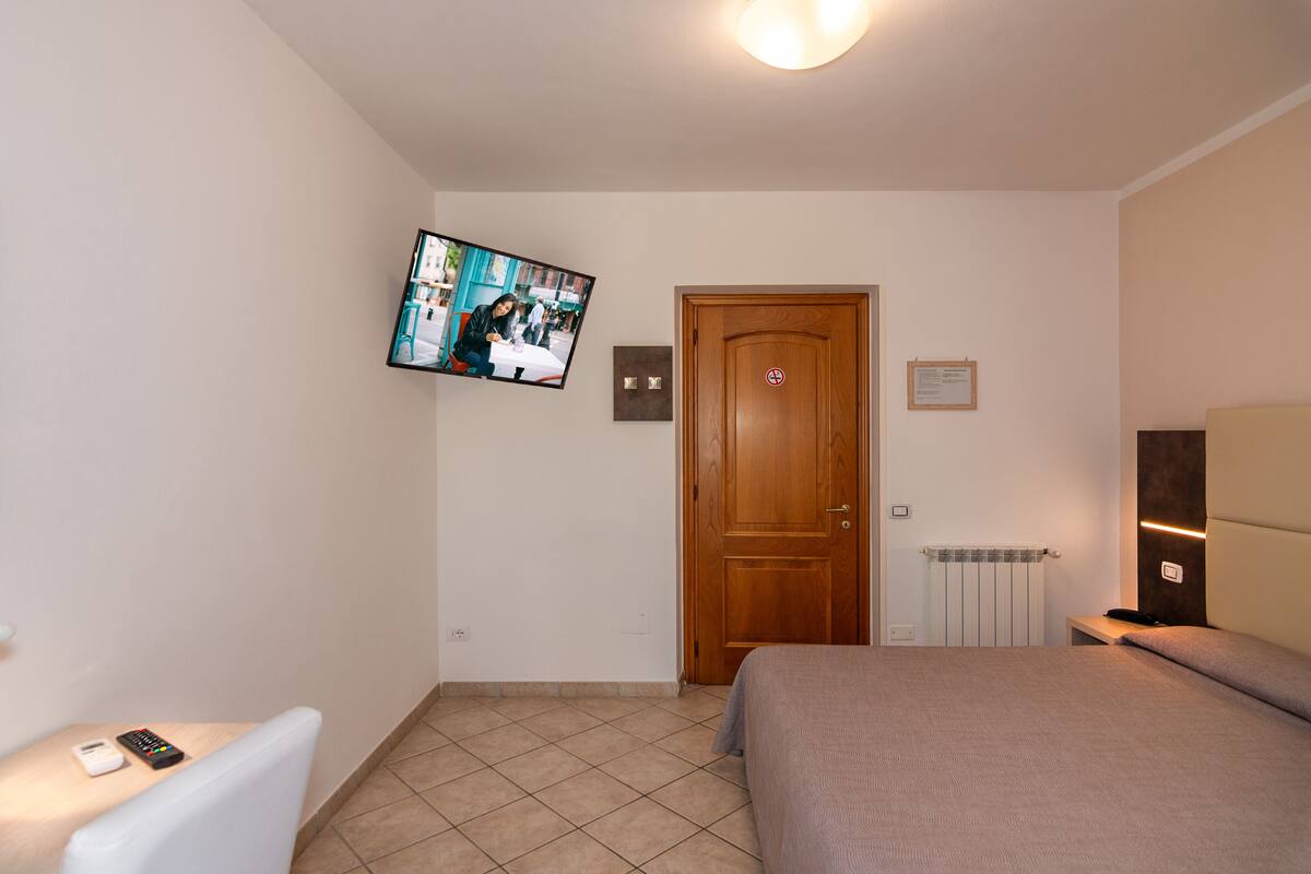 A hotel room with a bed, a TV mounted on the wall, a bedside table with a remote control, and a door with a sign.