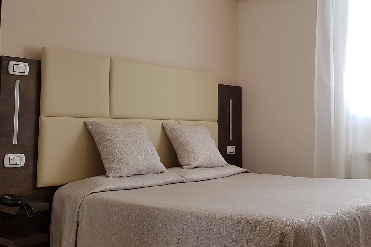 A hotel room with a bed, a desk with a remote control, and a window with sheer curtains.