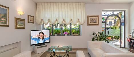 A living room with a television, white sofas, a glass coffee table, and a view of the street through a window with curtains.