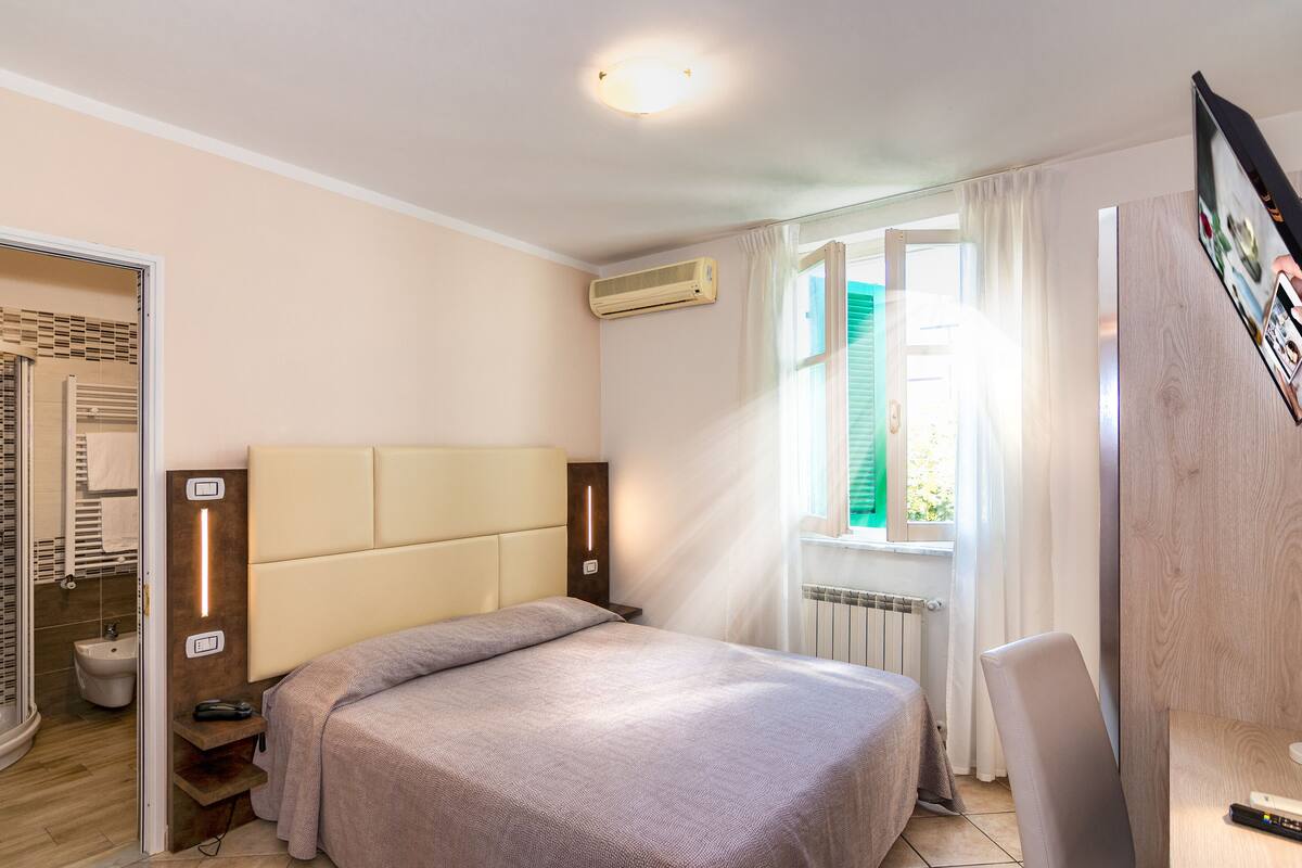 A hotel room with a bed, a bedside table, a chair, a bathroom with a shower, and a window with curtains.