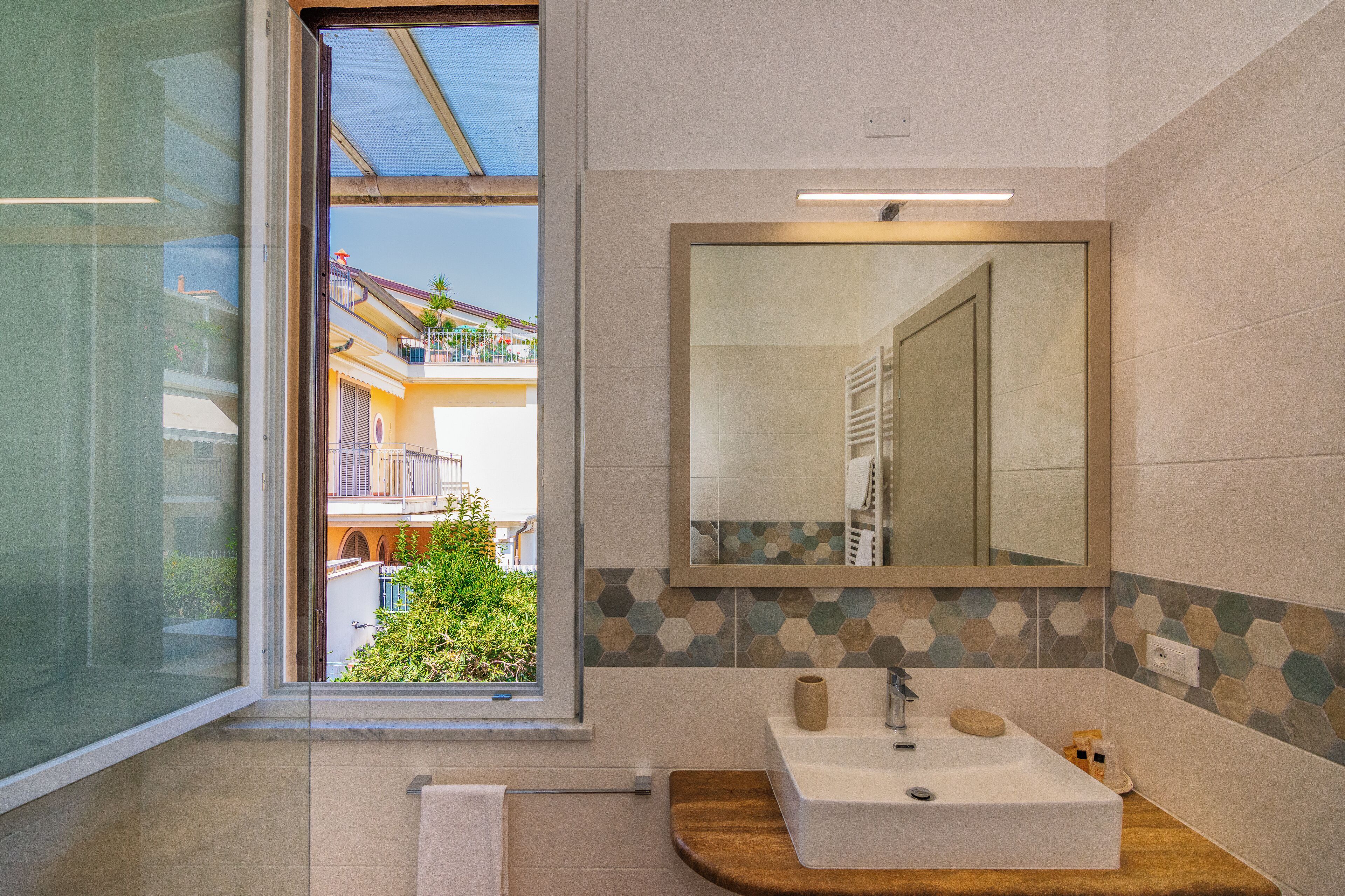Un baño moderno con un ventanal amplio, un mueble de tocador de madera, un lavabo blanco y un espejo con un revestimiento de azulejos hexagonales.
