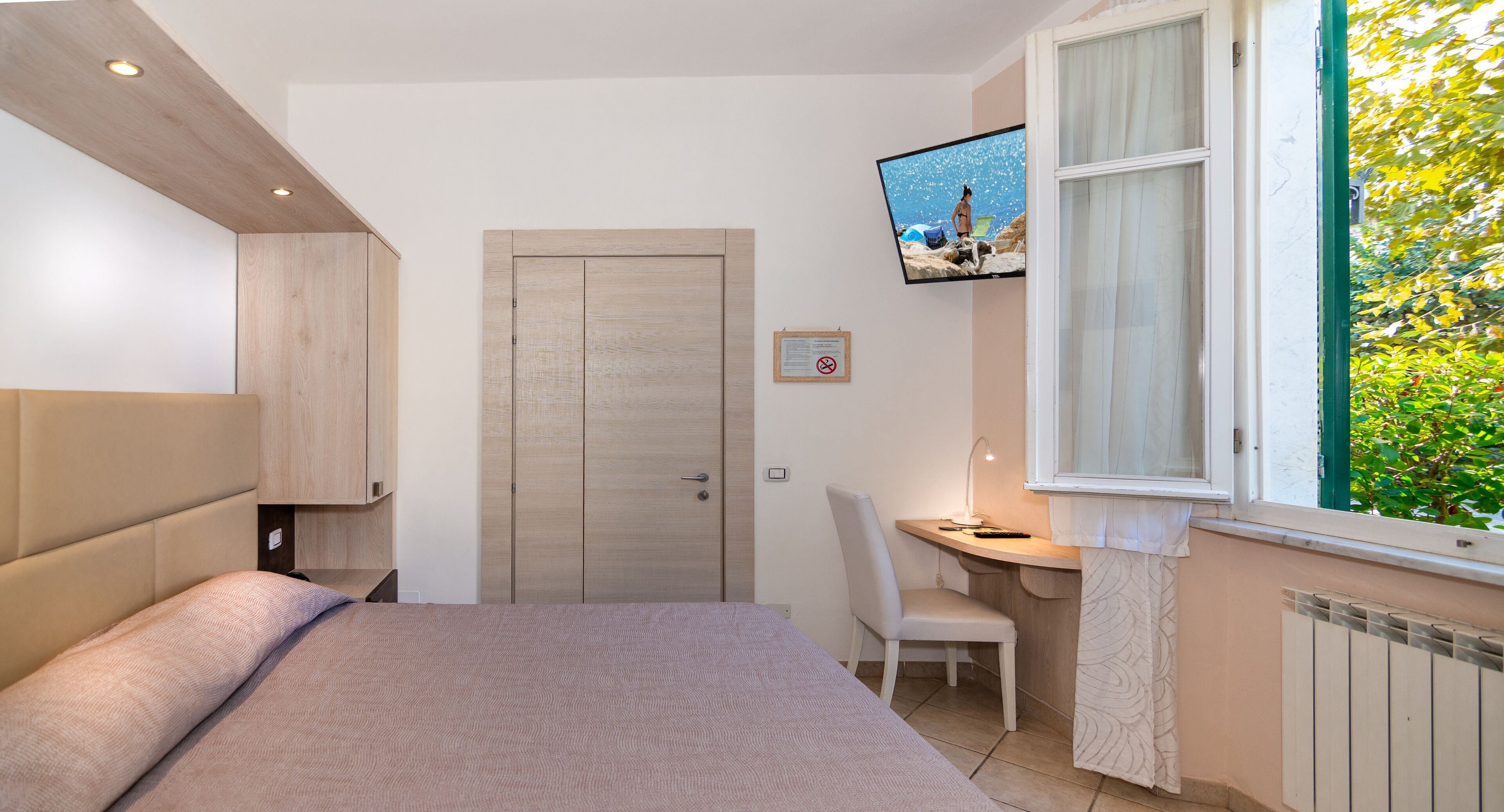 Un dormitorio con cama, escritorio, silla, televisor montado en la pared y ventana con vista a zonas verdes.