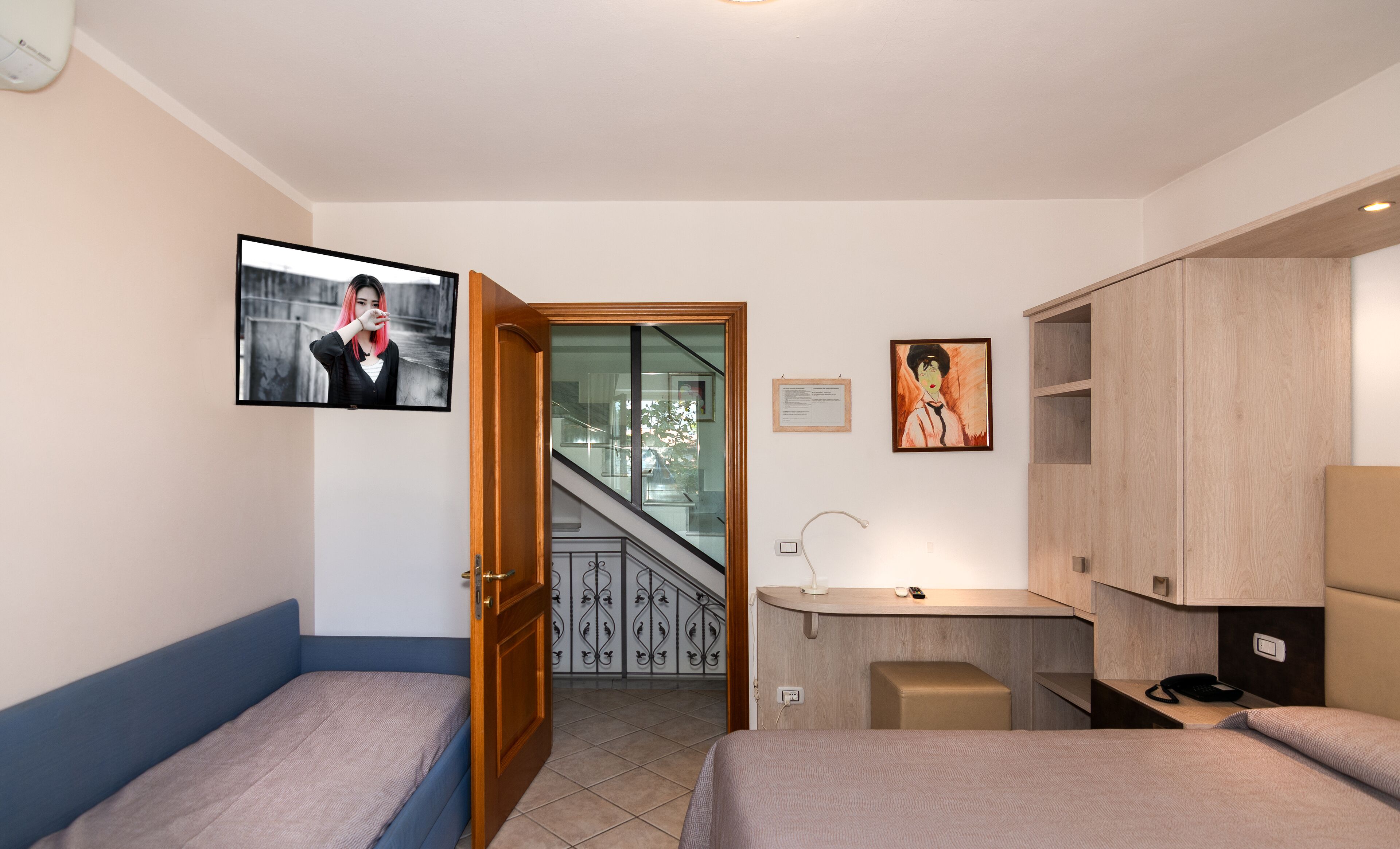 Habitación de hotel con dos camas individuales, un escritorio con una silla, un taburete pequeño, un televisor montado en la pared y una escalera que lleva a un área al aire libre.