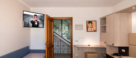 A hotel room with two single beds, a desk with a chair, a small stool, a TV mounted on the wall, and a staircase leading to an outdoor area.