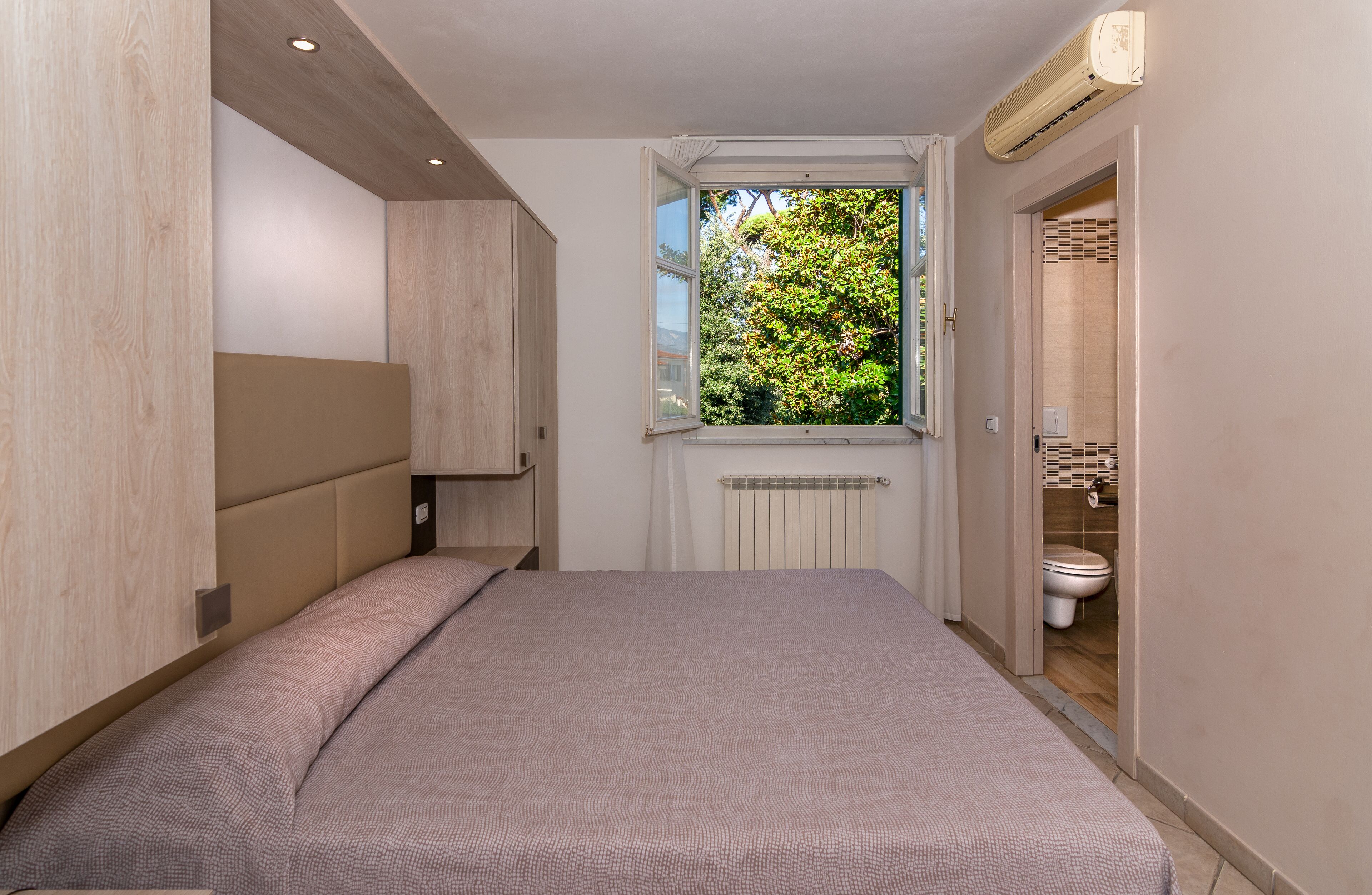 A bedroom with a bed, a window with a view of greenery, and a bathroom visible through an open door.