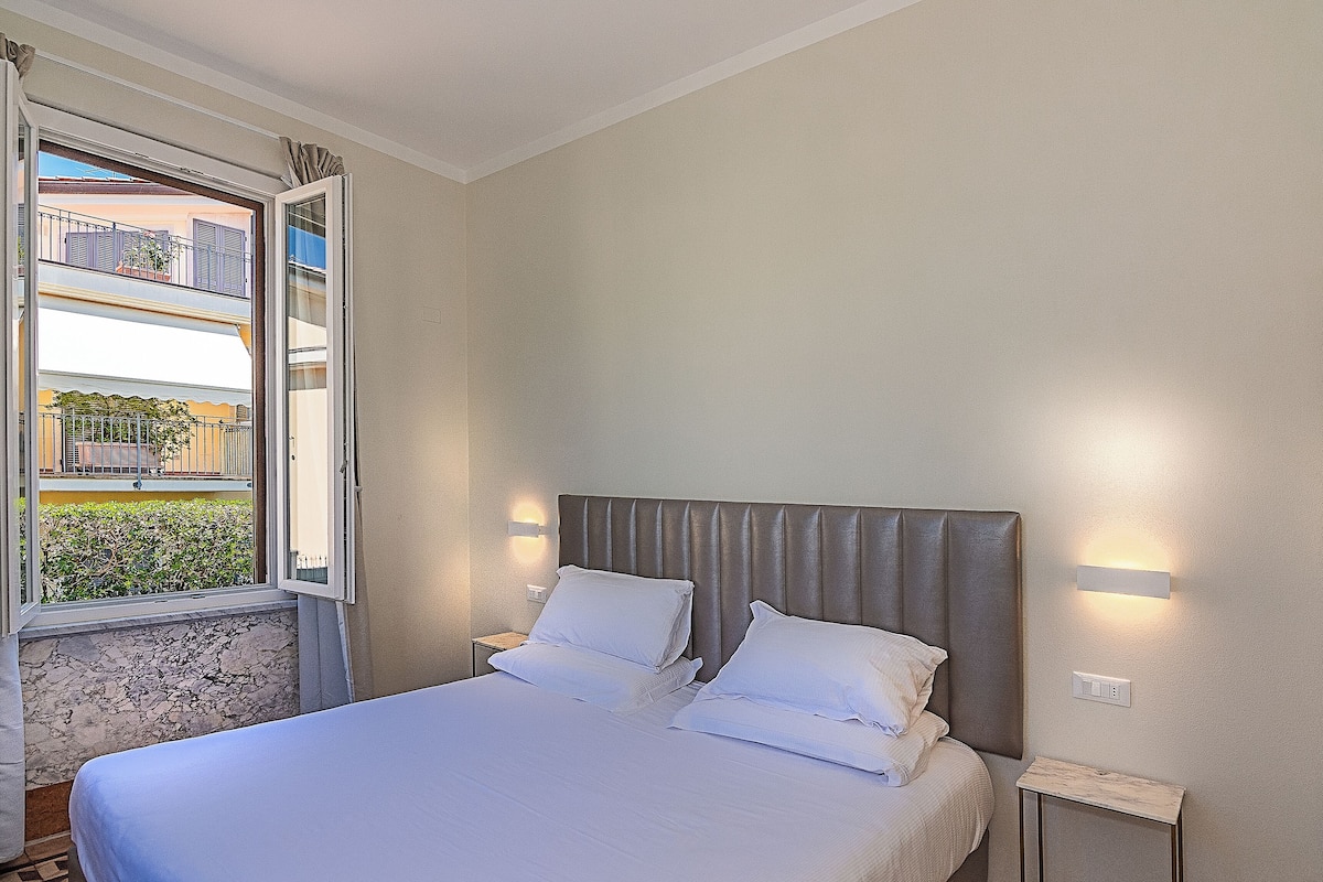 A bedroom with a bed, a nightstand, a wall-mounted light, and a window with a view of the outside.