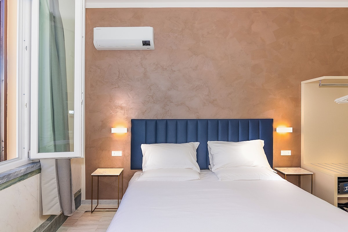 A hotel room with a large bed, a blue headboard, bedside lamps, an air conditioning unit, and a view of greenery outside.