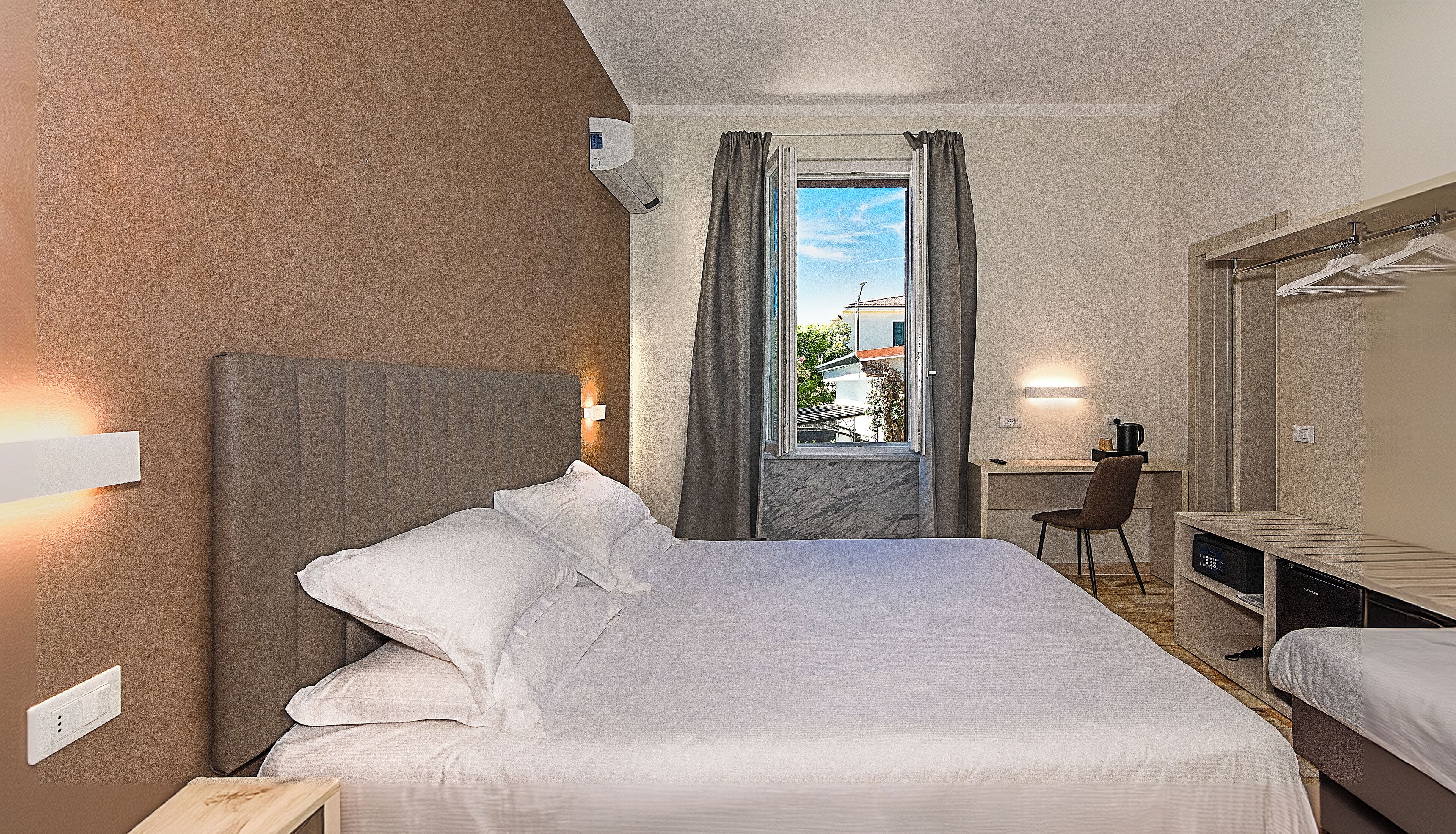 Habitación de hotel con cama, escritorio y silla. Se observa una ventana con cortinas, un aire acondicionado de pared y una lámpara de pared.