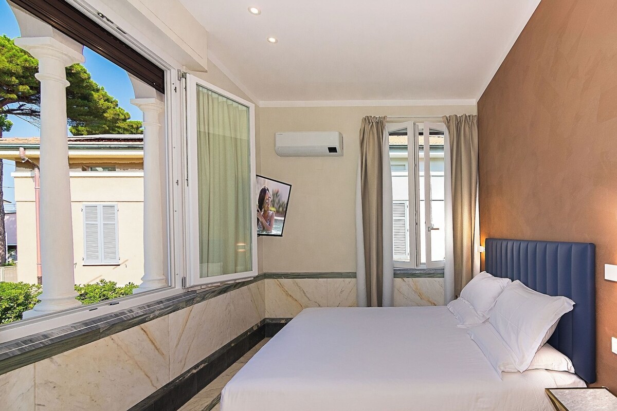 A hotel room with a bed, a window with curtains, a wall-mounted air conditioner, and a picture on the wall.