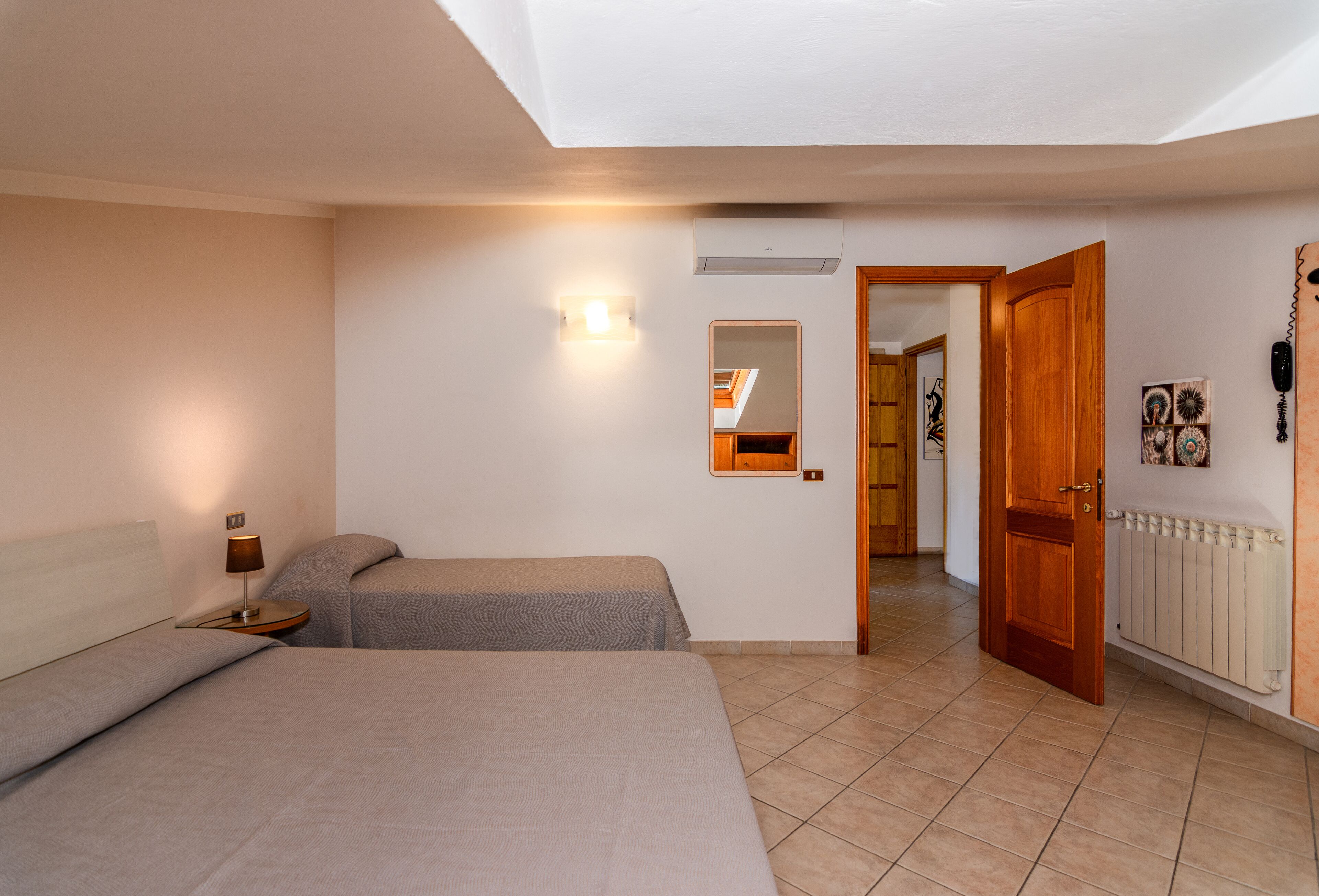A bedroom with two beds, a bedside table with a lamp, a wall-mounted light, a mirror, and a door leading to another room.