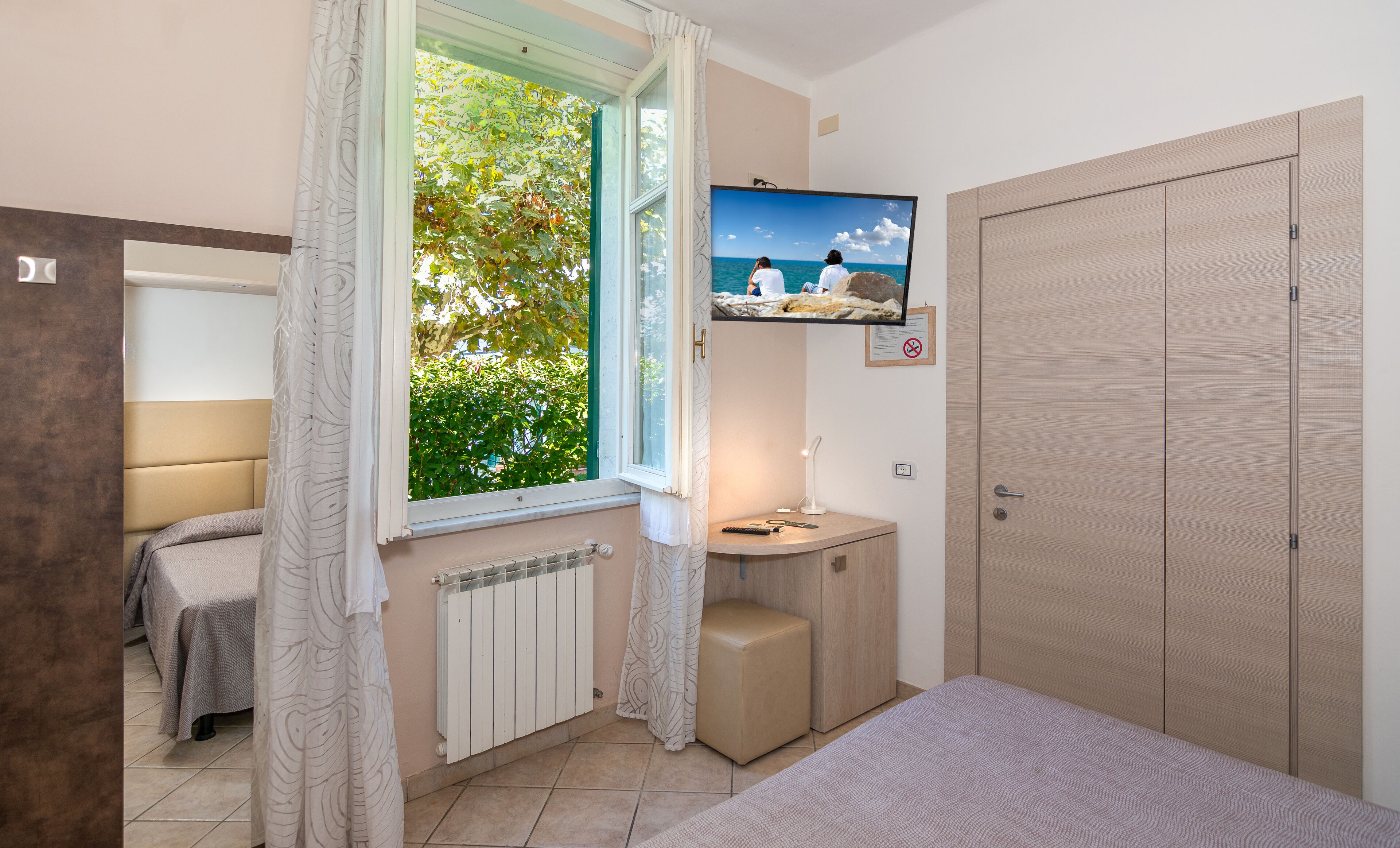 A bedroom with a bed, a window with curtains, a TV on the wall, a bedside table, and a wardrobe.
