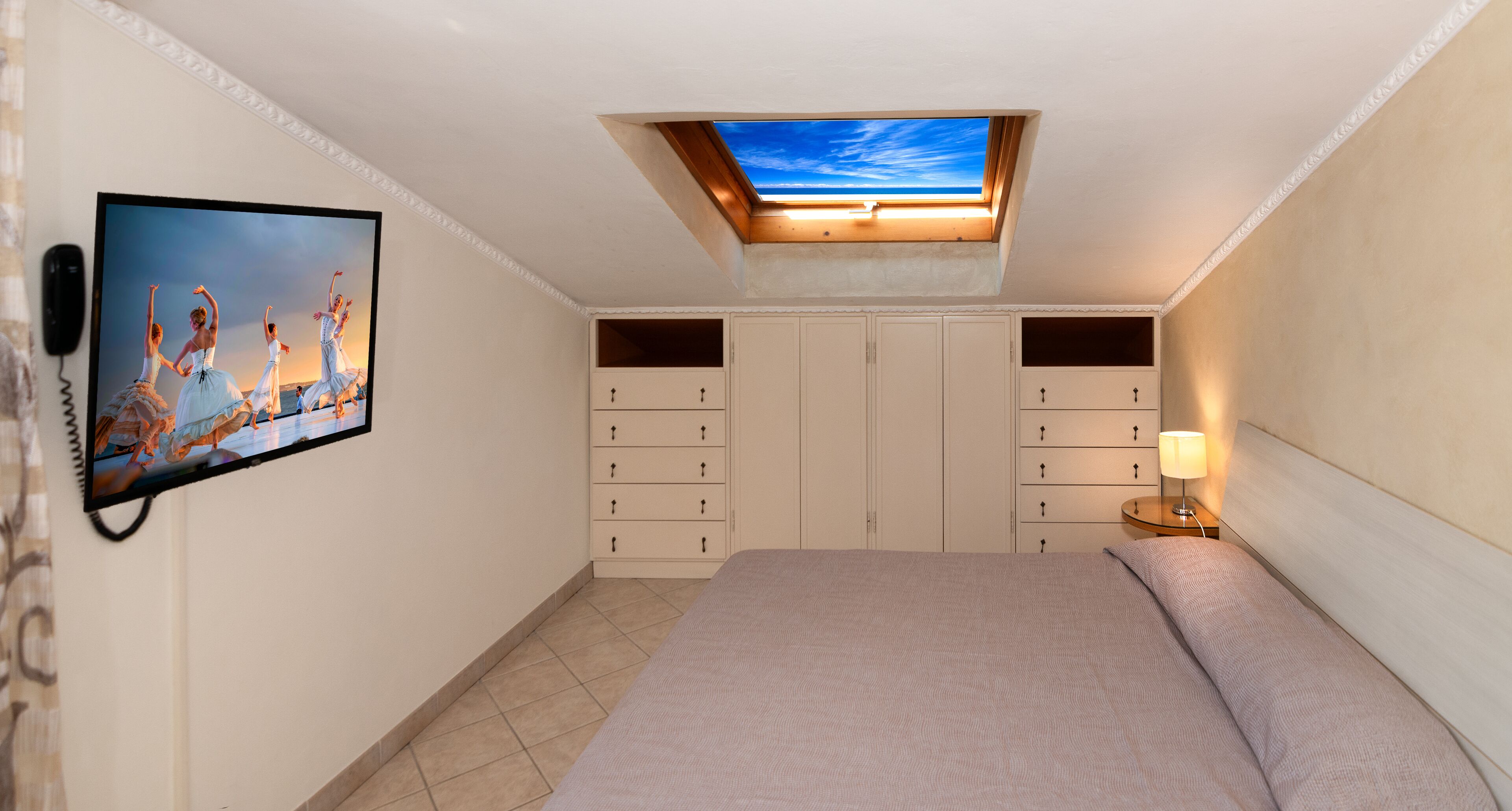 A bedroom with a bed, a TV mounted on the wall, a desk with a lamp, and a wardrobe.