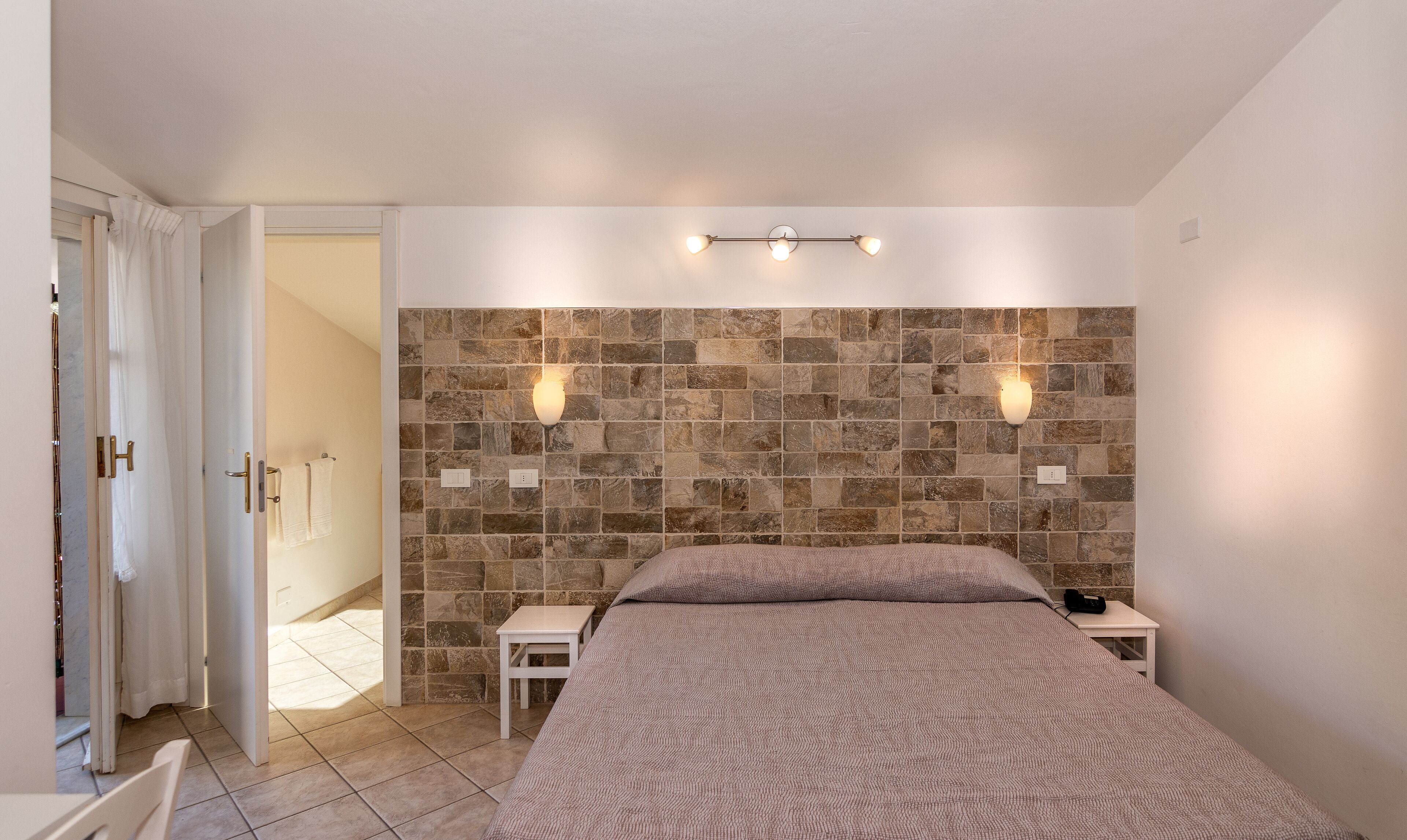 Un dormitorio con cabecera de pared de piedra, dos lámparas de noche, una cama con manta texturizada y una mesita de noche pequeña.