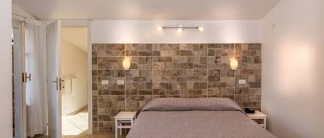 Een slaapkamer met een hoofdeinde van natuursteen, twee bedlampen, een bed met een gebreid plaid en een klein nachtkastje.