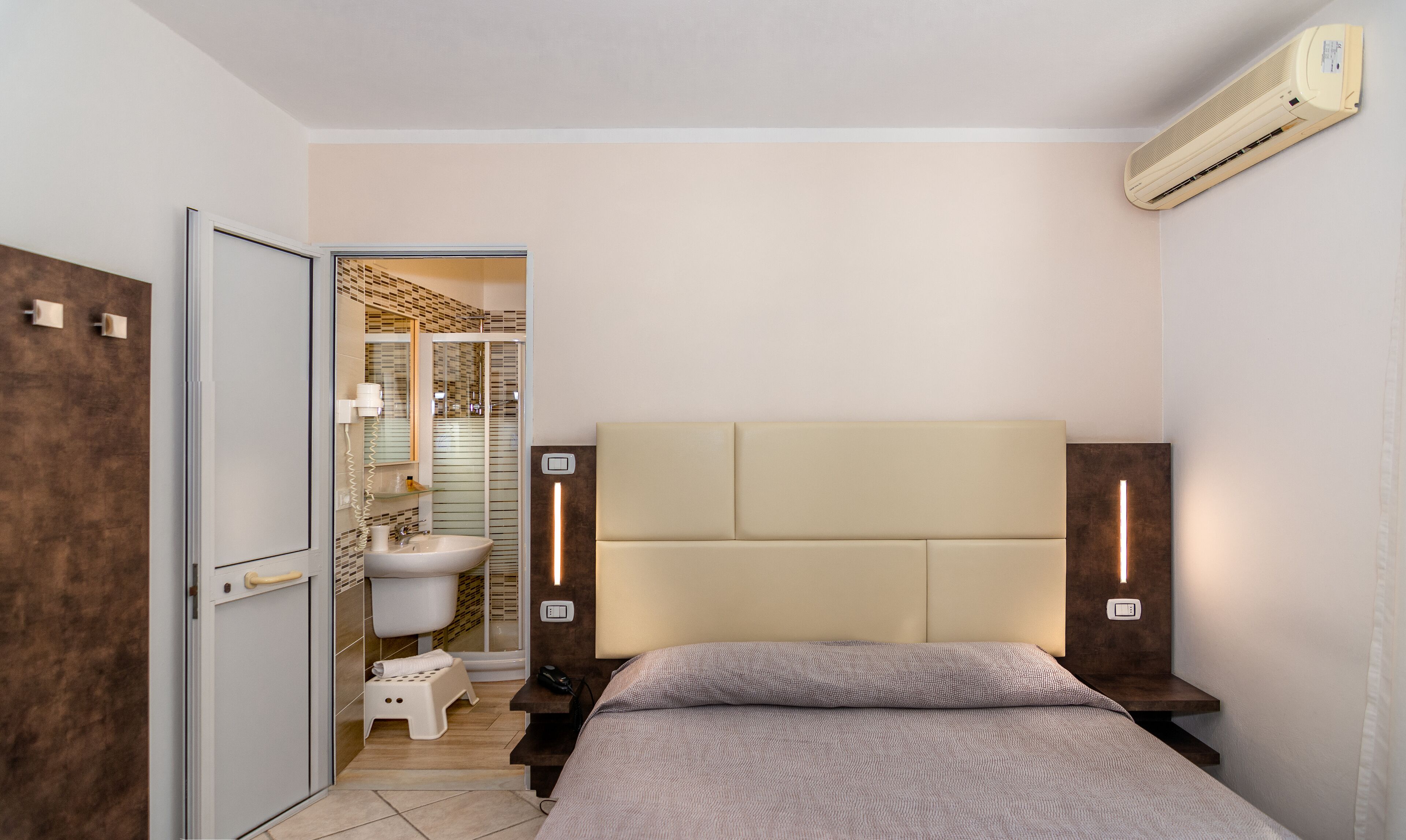 Une chambre à coucher comprenant un lit, des tables de chevet, un appareil de climatisation, une salle de bain avec des toilettes, une douche et un porte-serviettes.