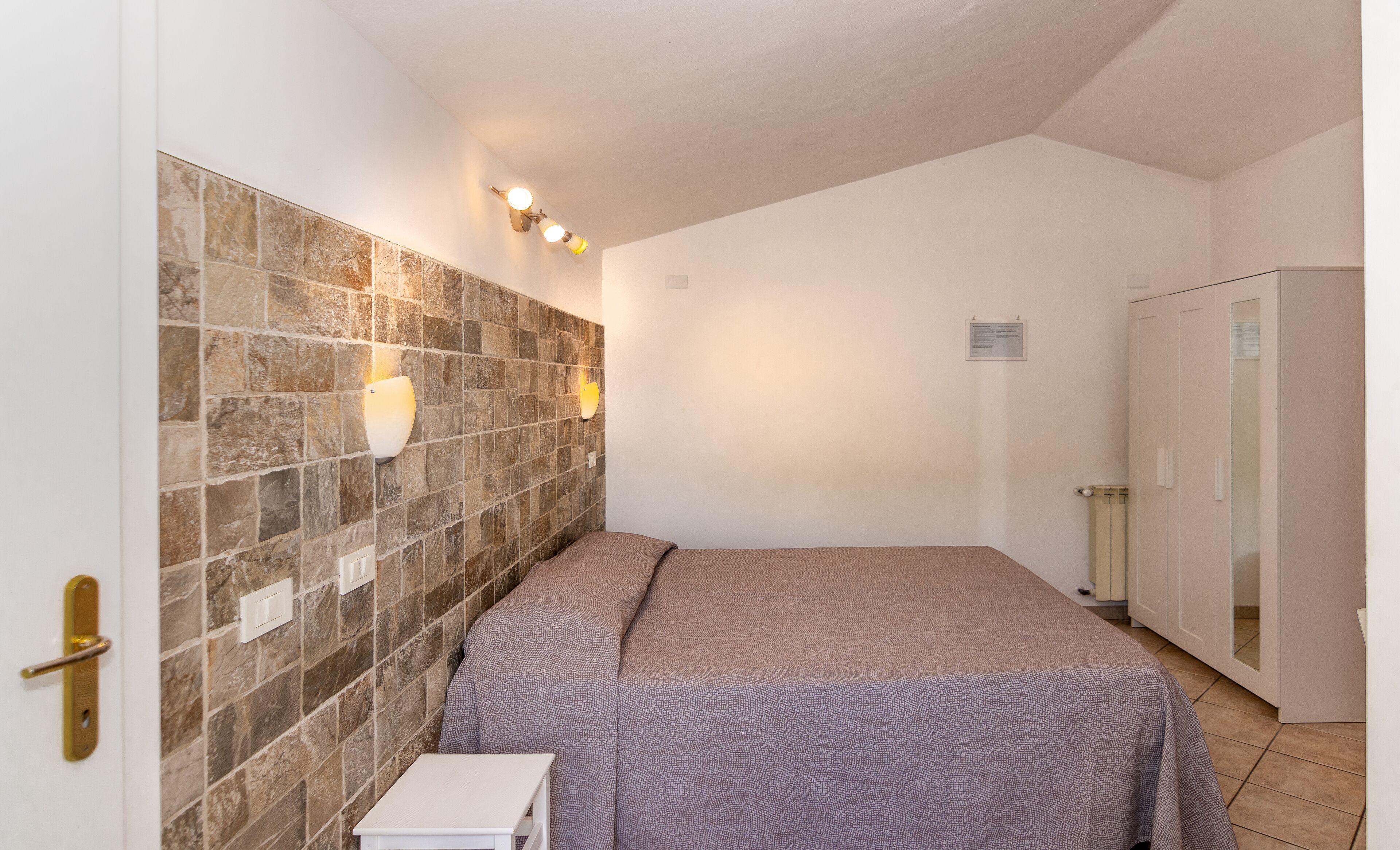 A bedroom with a stone-patterned wall, a bed with patterned bedding, a small bedside table, and a wardrobe.