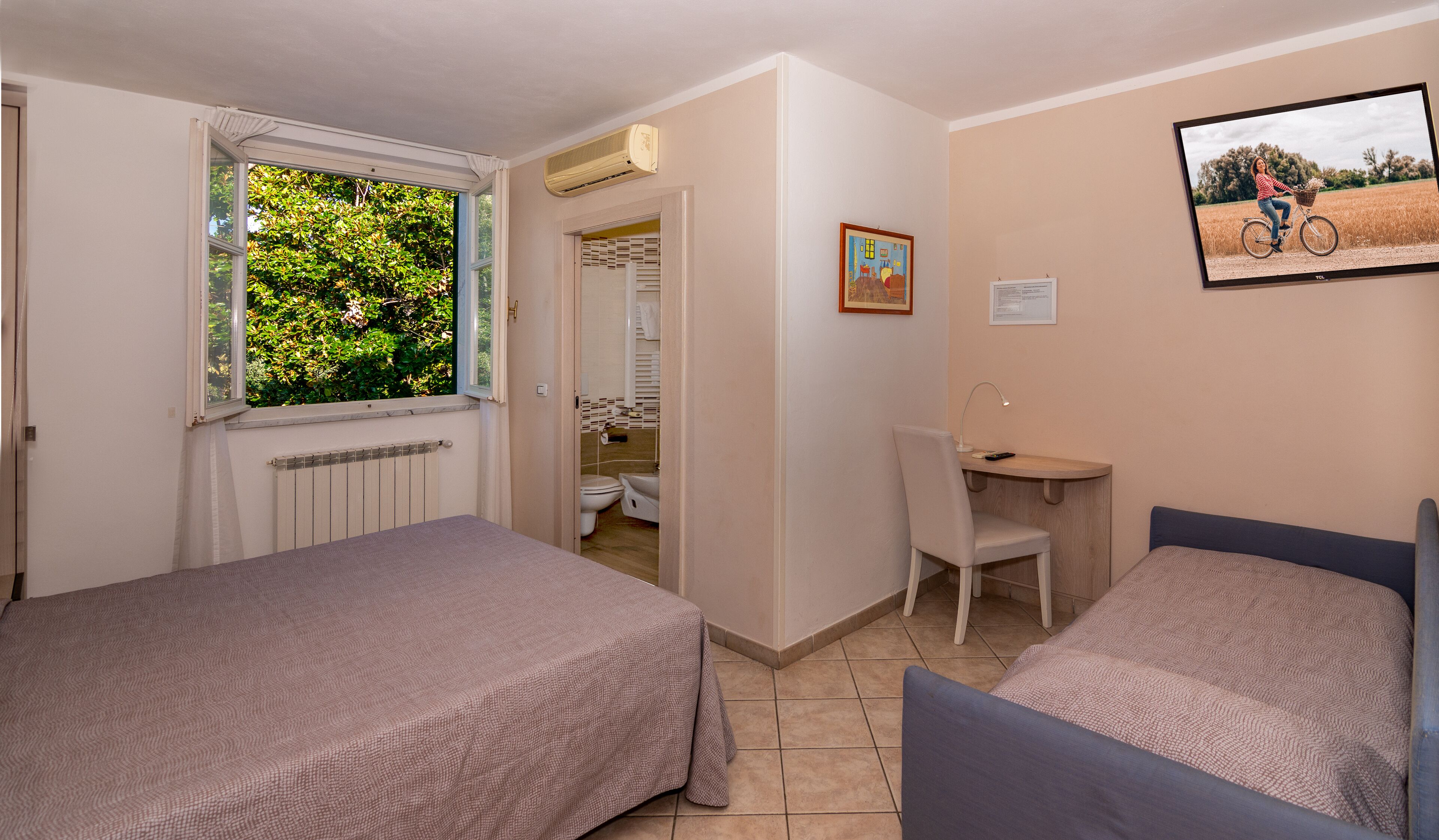 Een slaapkamer met twee eenpersoonsbedden, een bureau, een stoel, een radiator, een raam met uitzicht op groen en een lijst met een afbeelding aan de muur.