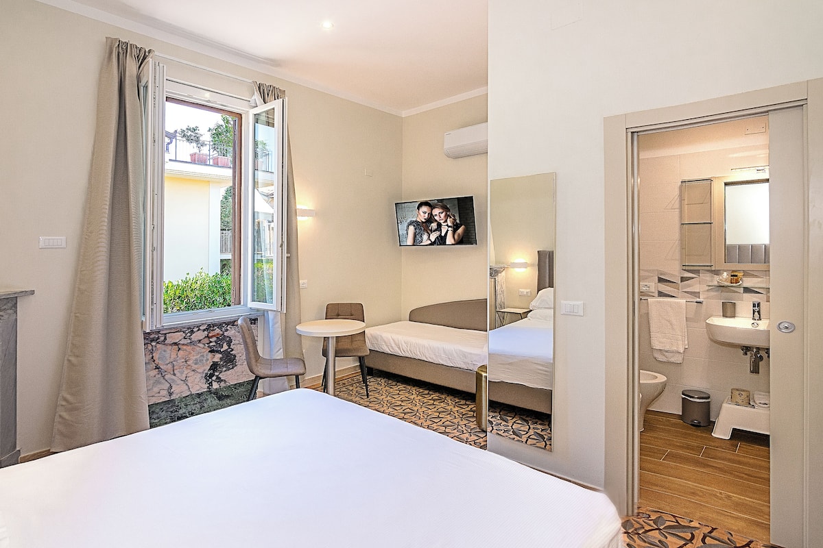A hotel room with a bed, a small table, a chair, a TV, and a bathroom visible through an open door.
