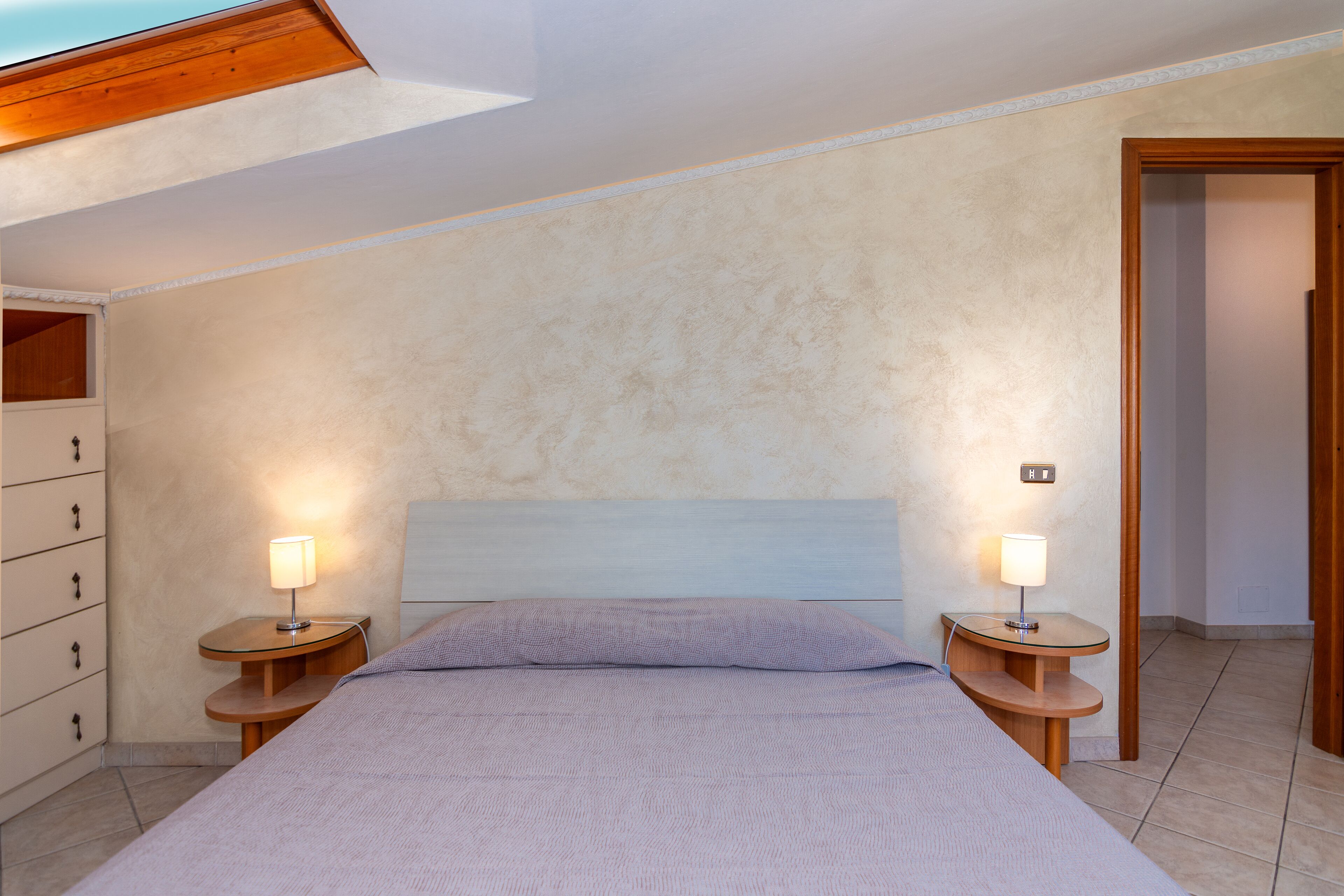 A bedroom with a bed, two bedside tables with lamps, a wooden dresser, and a door.