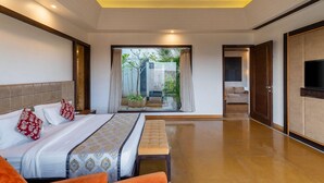 A modern hotel room with a large bed, a bench, a TV, and a view of the garden through a glass door. - Mahua Bagh Resort (Kumbhalgarh)