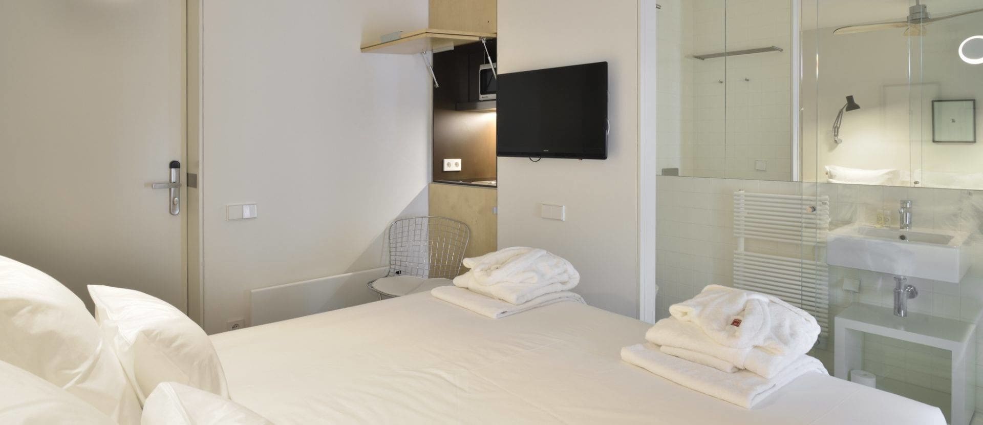 Une chambre d’hôtel avec un lit, un téléviseur fixé au mur, une petite table et une salle de bain équipée d’un lavabo et d’un miroir.