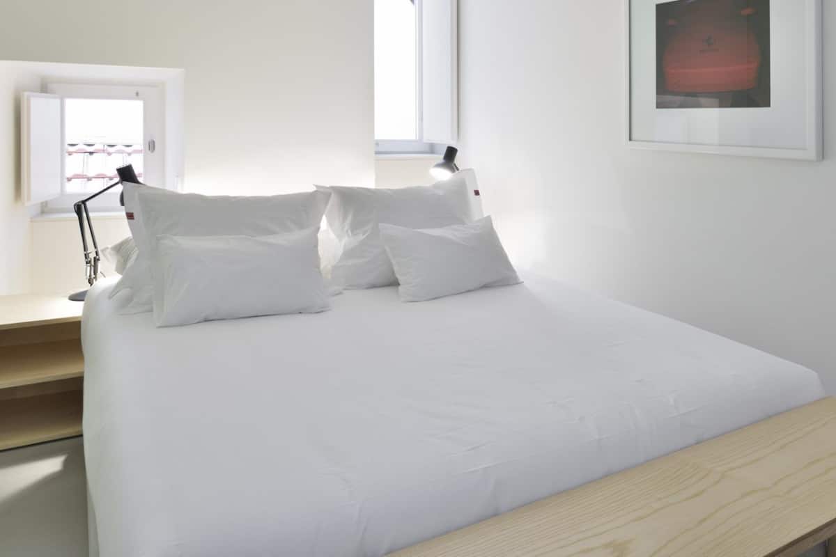 A neatly made bed with white linens, a bedside table with a lamp, and a framed picture on the wall.