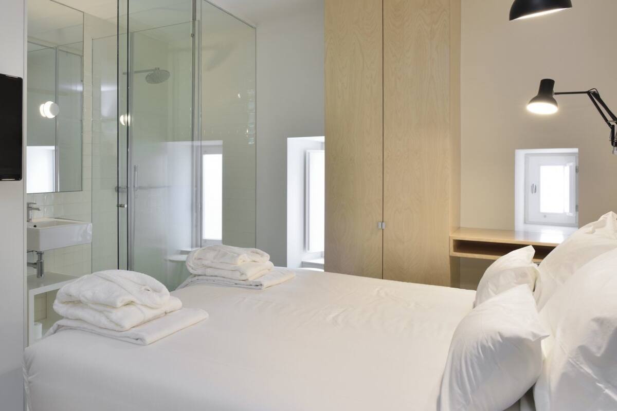 A modern hotel room with a bed, a TV mounted on the wall, a glass shower enclosure, and a bedside lamp.