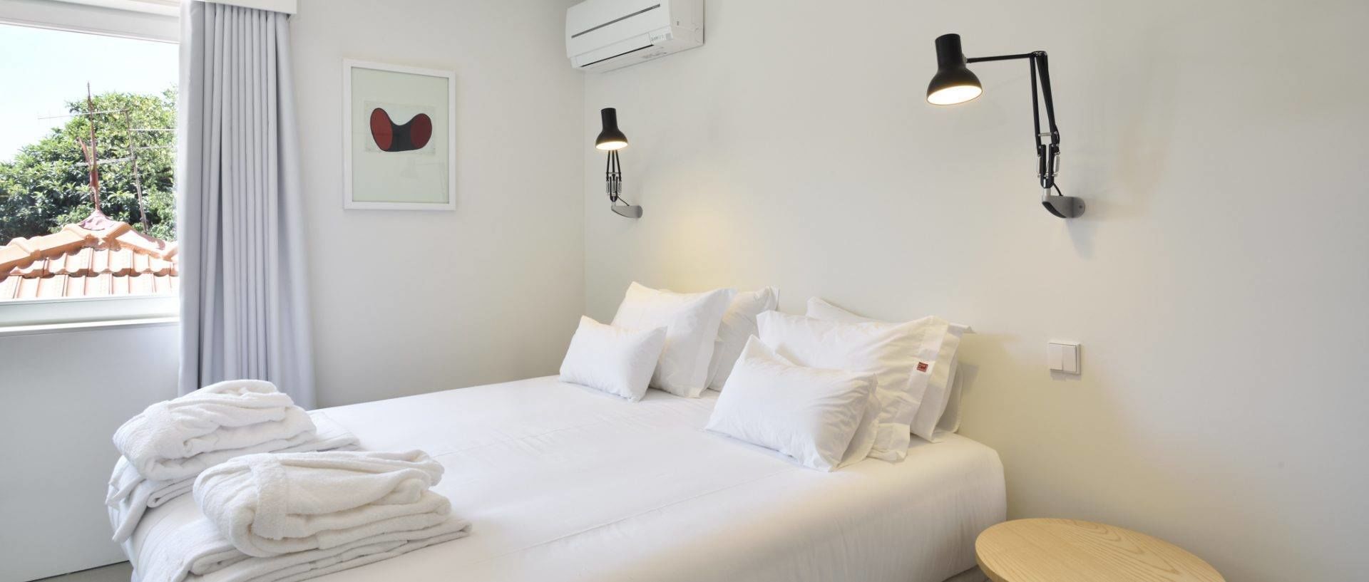 A neatly made bed with white linens and pillows, a stack of folded towels, and two wall-mounted reading lamps.