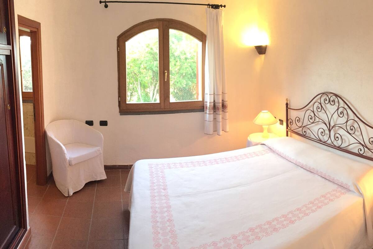 Ein Schlafzimmer mit einem Bett, zwei Nachttischlampen, einem Fenster mit Blick auf Grünflächen, einem Sessel und einer Holztür.