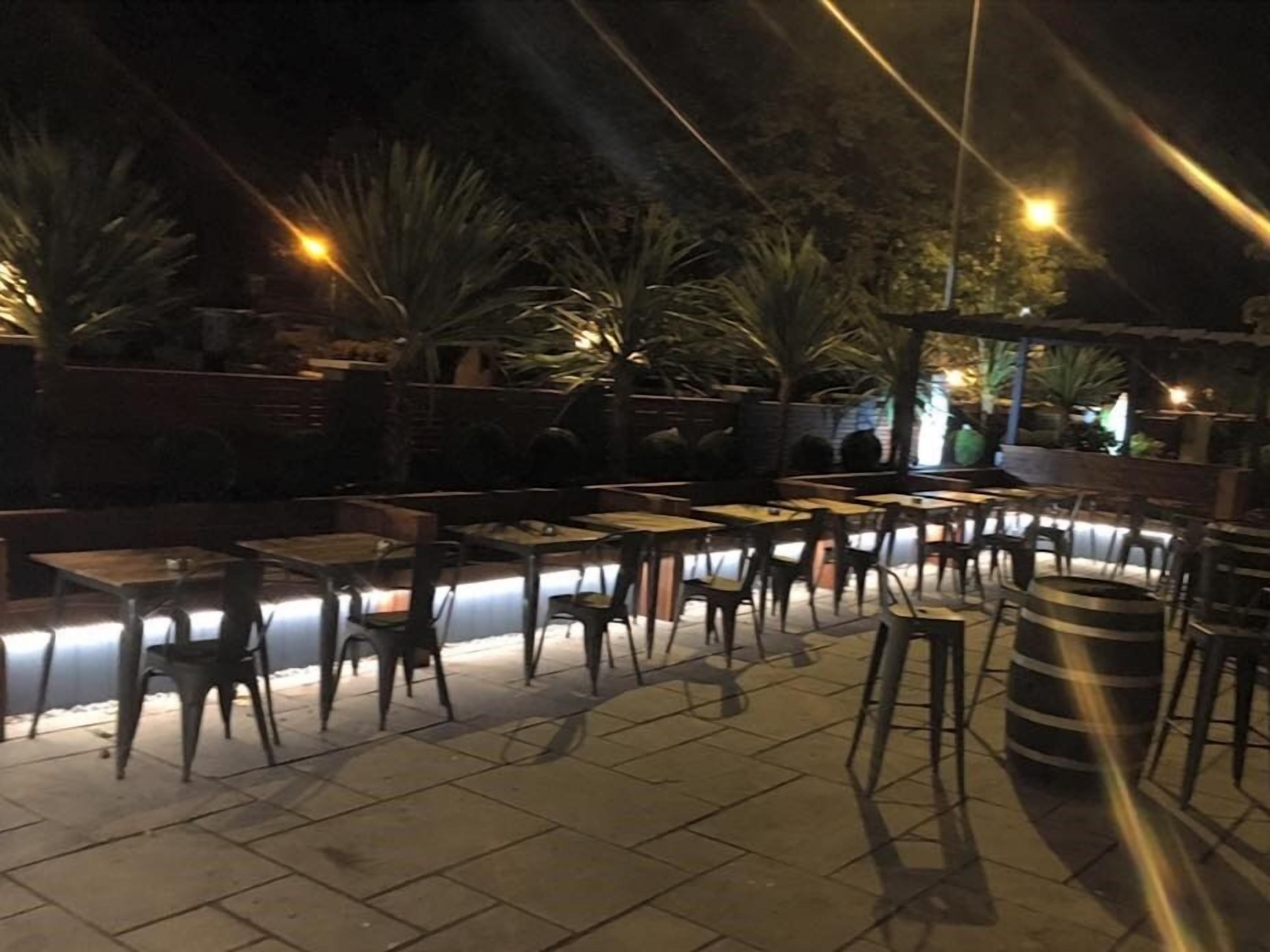 An outdoor patio area with tables and chairs, surrounded by plants and a stone floor.