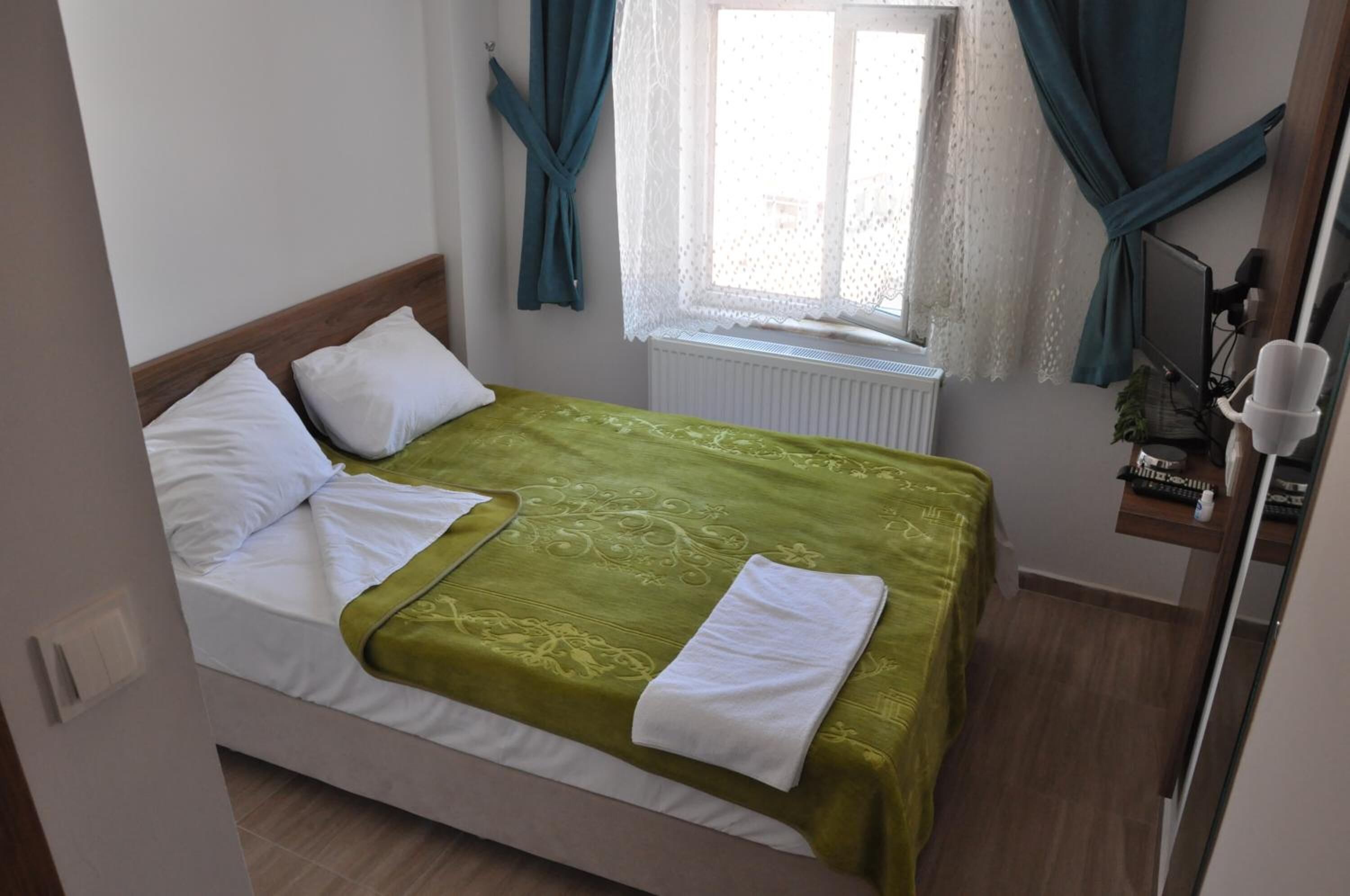 Una cama bien hecha con colcha verde y almohadas blancas, una ventana con cortinas, un radiador, un estante con objetos y un espejo.