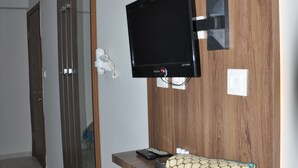 Quarto de hotel com parede de painel de madeira, televisão montada, uma prateleira pequena com um objeto decorativo e um espelho.