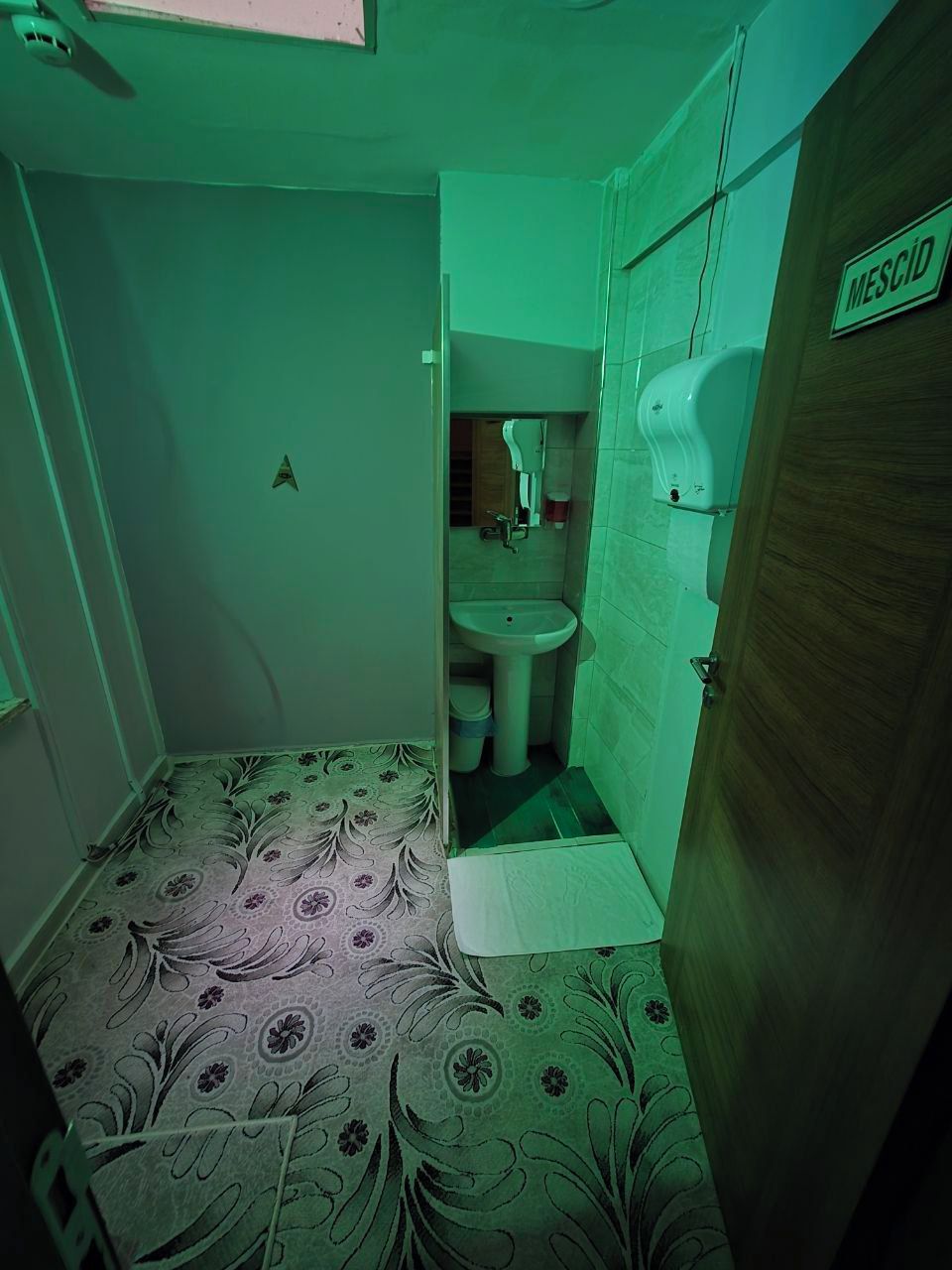 Un baño pequeño con lavamanos, inodoro y zona de ducha. Un cartel en la pared dice “MESCID”.
