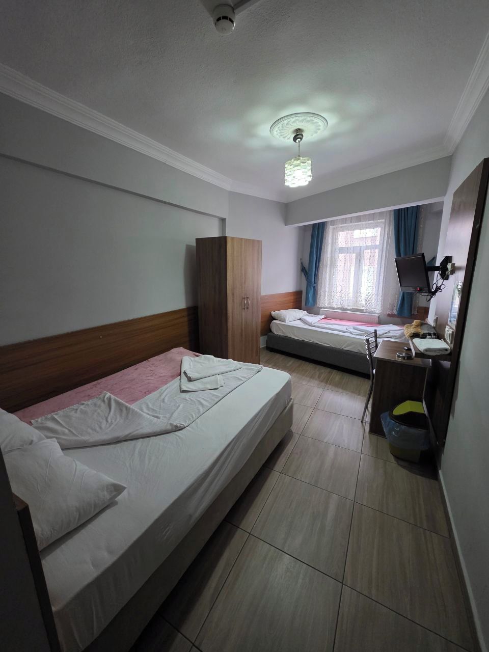 A hotel room with two beds, a desk, a wardrobe, a ceiling fan, and a window with curtains.