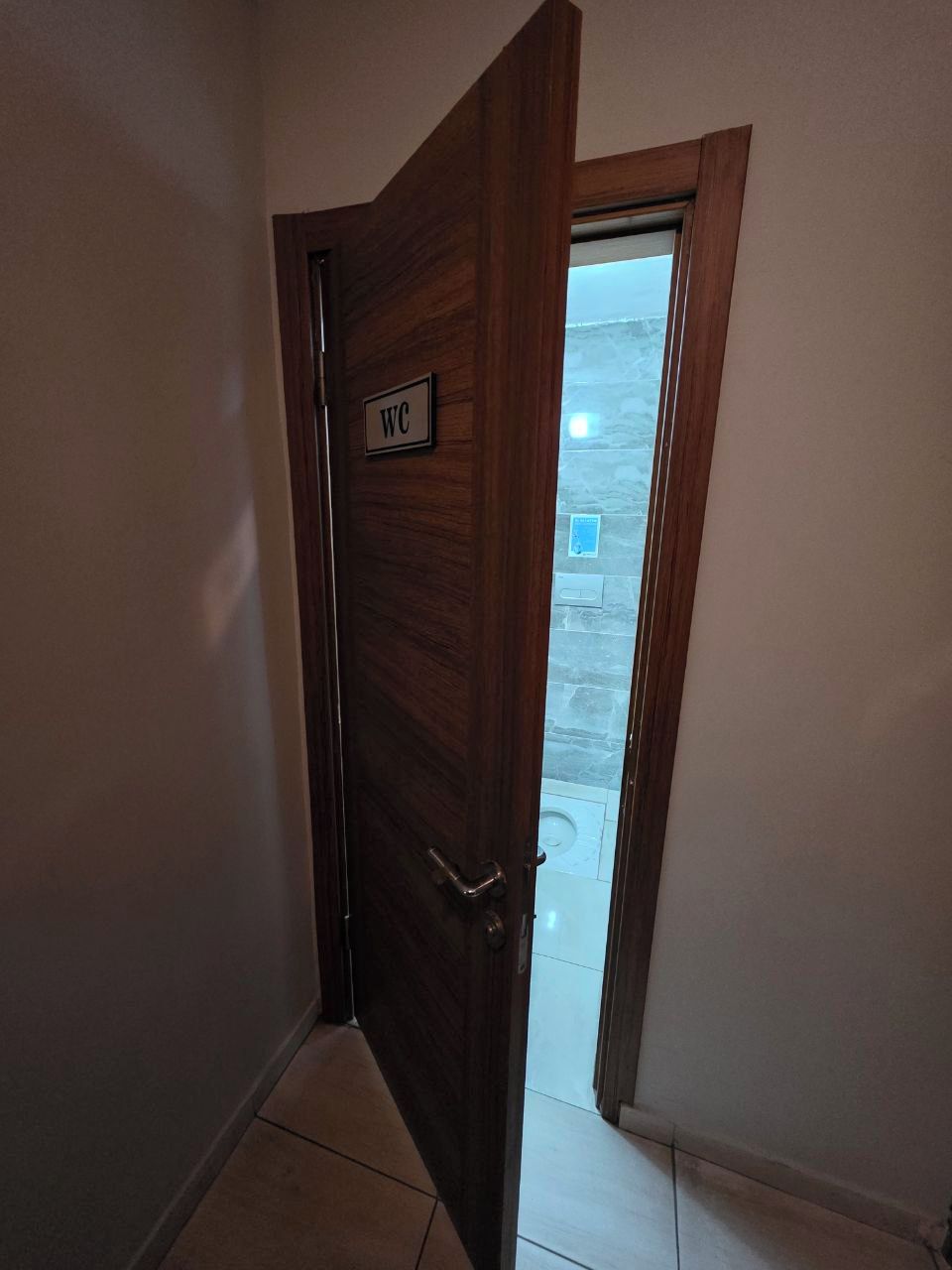 Una puerta de madera con el símbolo de WC, que da a un espacio exterior con un muro de piedra y suelo de baldosas.