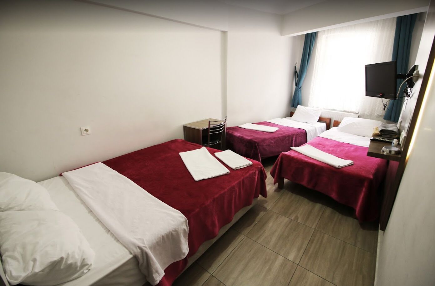 Quarto de hotel com duas camas de solteiro, uma escrivaninha, uma cadeira, televisão e janela com cortinas.