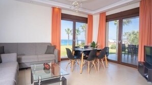 A modern living room with a sofa, glass coffee table, dining table with chairs, and a TV. Large windows with curtains offer a view of the outdoors. - Belohorizonte Fine Accommodation (Kassandra)