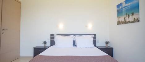 A bedroom with a bed, two bedside tables, a nightstand, a door, and a wall-mounted light fixture.