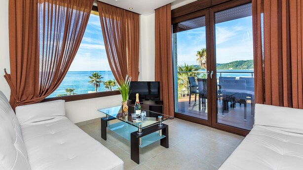 A modern living room with a glass coffee table, white sofa, and a TV. Large windows with brown curtains offer a view of the sea and palm trees. - Belohorizonte Fine Accommodation (Kassandra)
