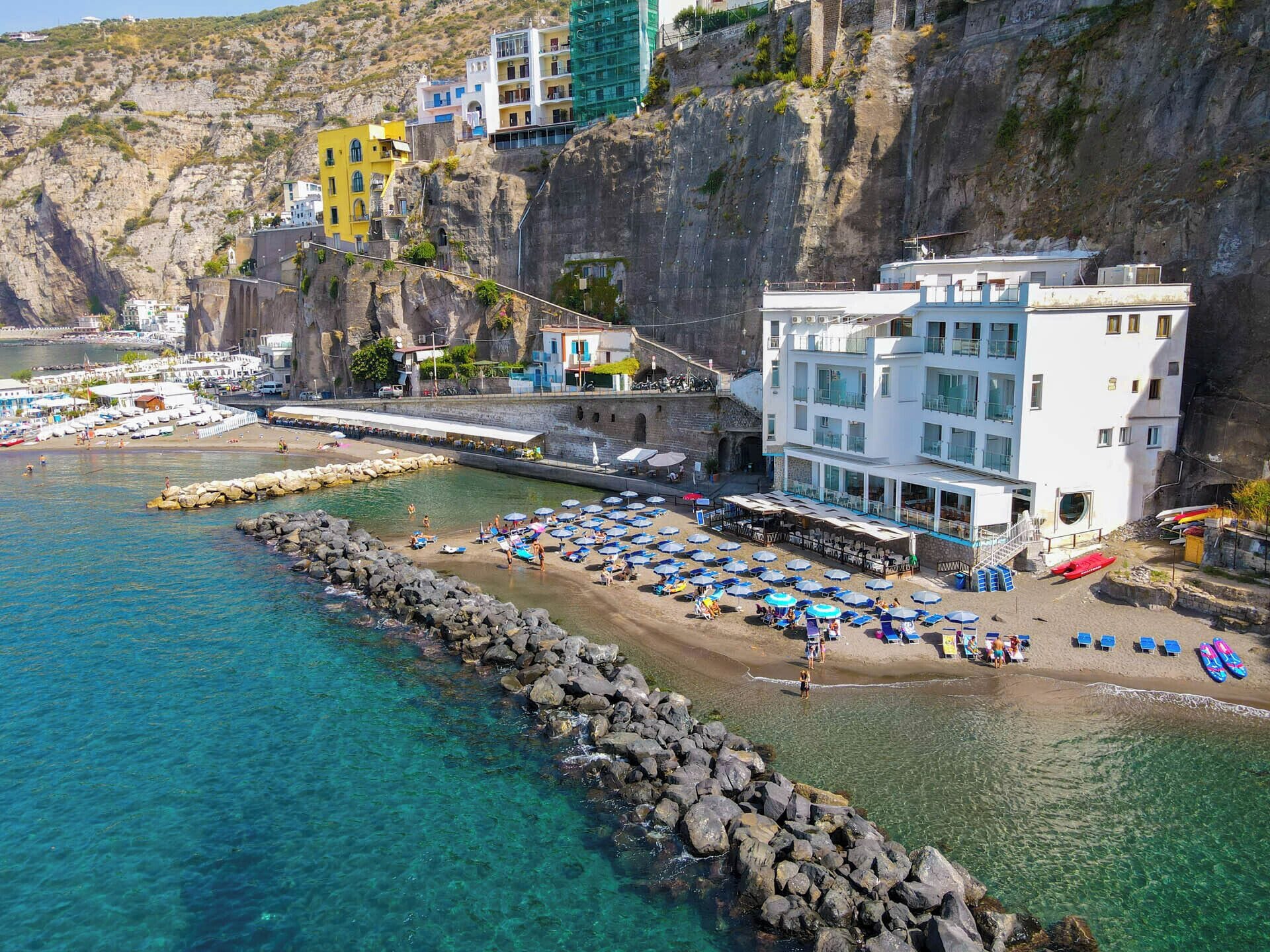 A coastal hotel with a rocky shoreline, clear blue waters, and a beach area with umbrellas and sun loungers.