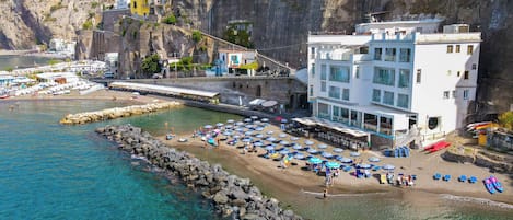 A coastal hotel with a rocky shoreline, clear blue waters, and a beach area with umbrellas and sun loungers.