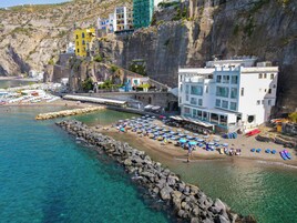 Un hotel frente al mar con costa rocosa, aguas azul cristalino y una zona de playa con sombrillas y tumbonas.
