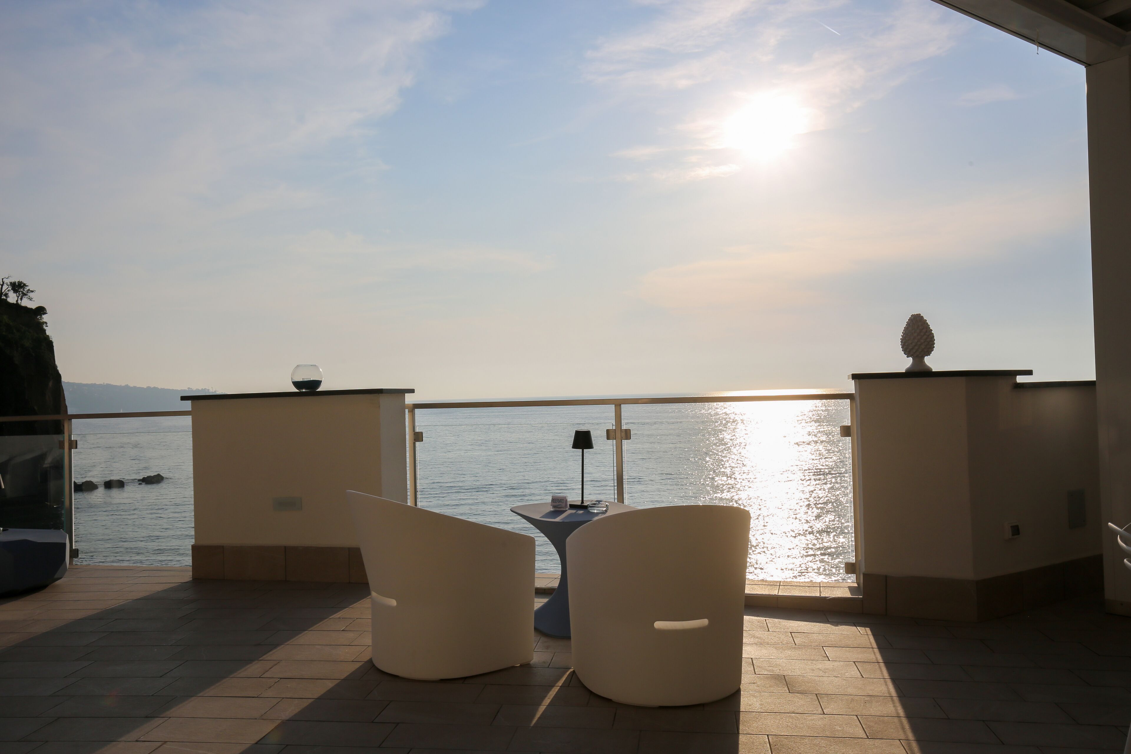 A terrace with two white chairs and a small table, overlooking a body of water with a clear sky and the sun setting or rising.