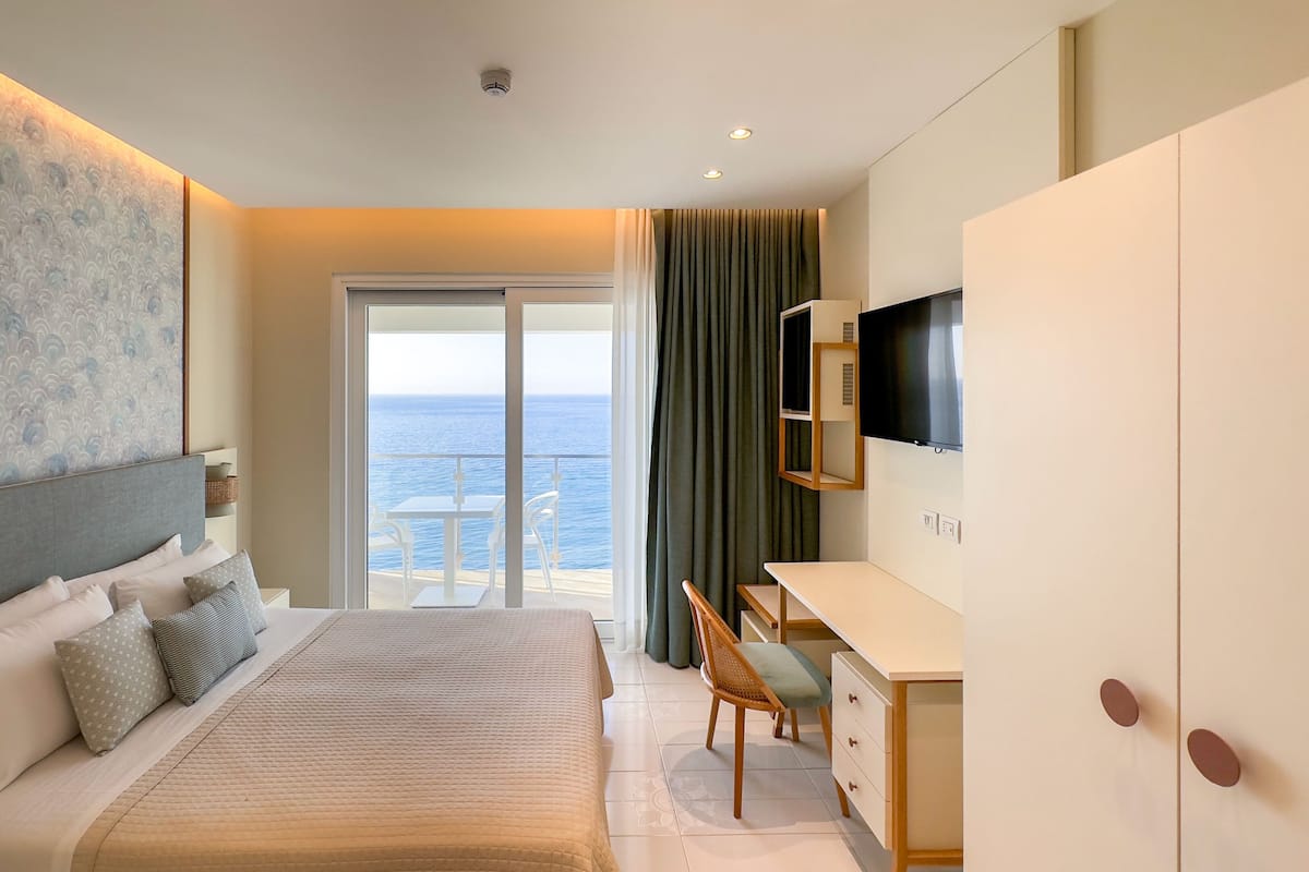 Habitación de hotel con una cama grande, un escritorio, un televisor y un balcón con vista al mar.