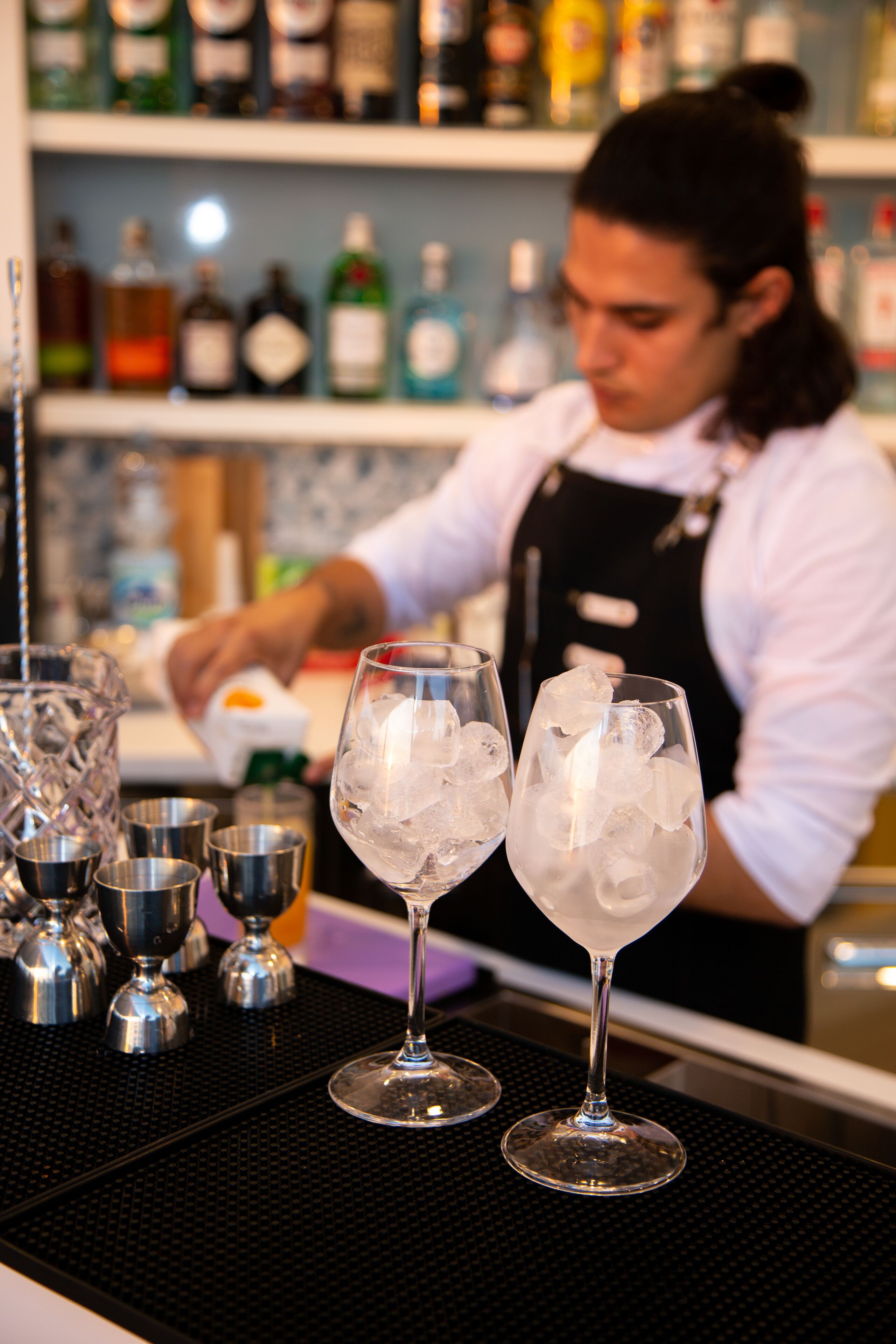 Un bar avec deux verres d’eau glacée, un barista préparant des boissons et, au fond, des étagères remplies de différentes bouteilles.