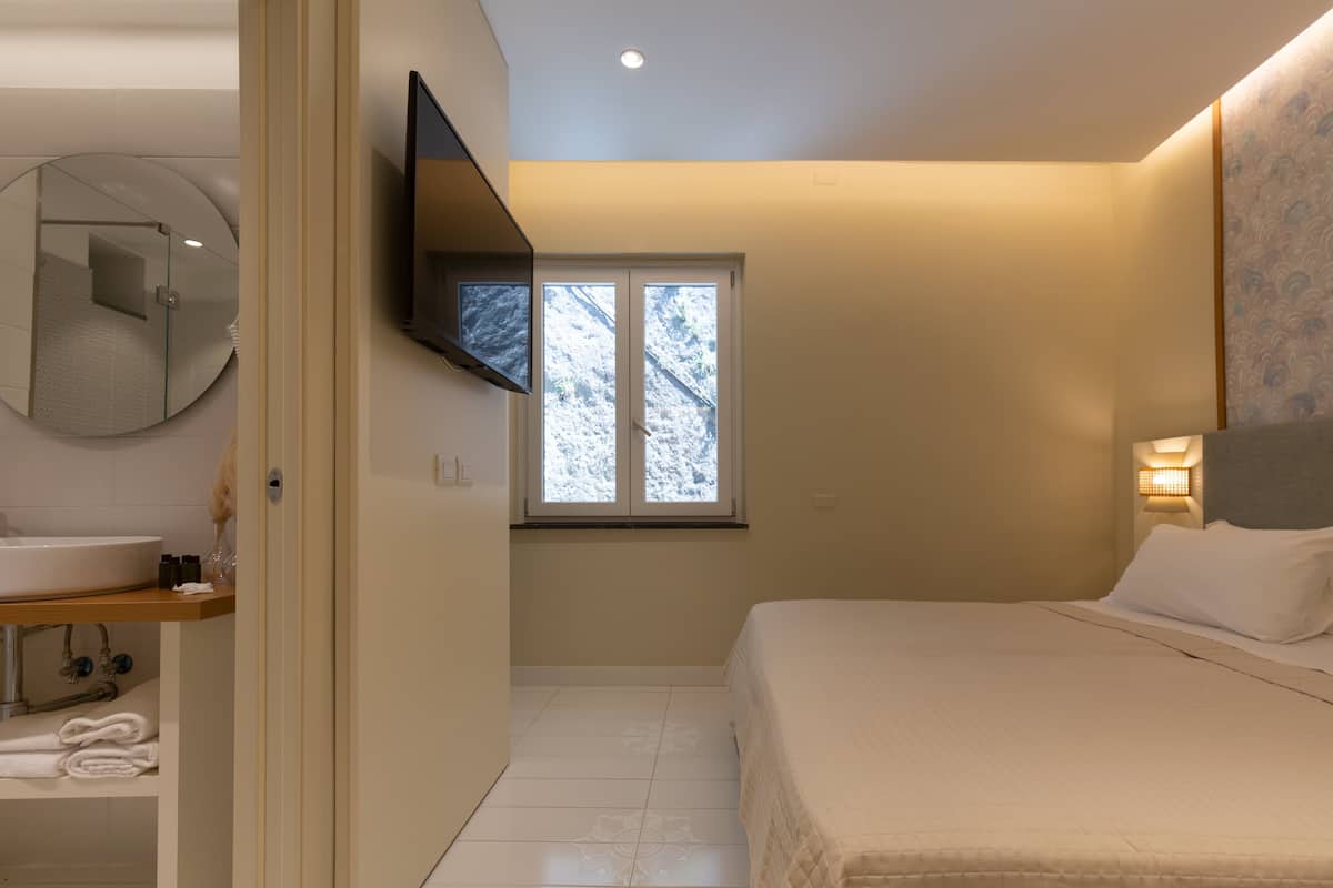 Un dormitorio con cama, televisor montado en la pared, ventana con vistas a árboles y baño con lavabo y espejo.