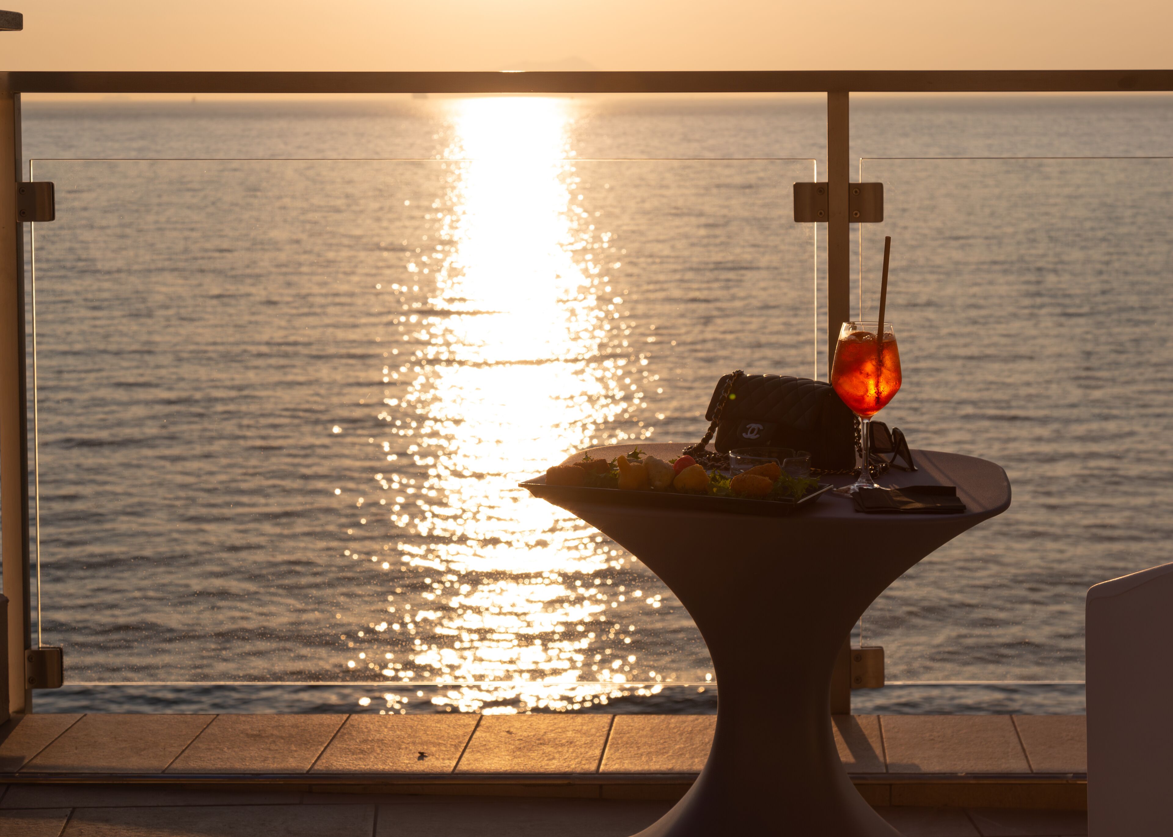 A table with a glass of drink and a plate of food, set against a view of the sea at sunset.