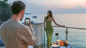 Un hombre fotografiando a una mujer en un balcón con vista al mar.