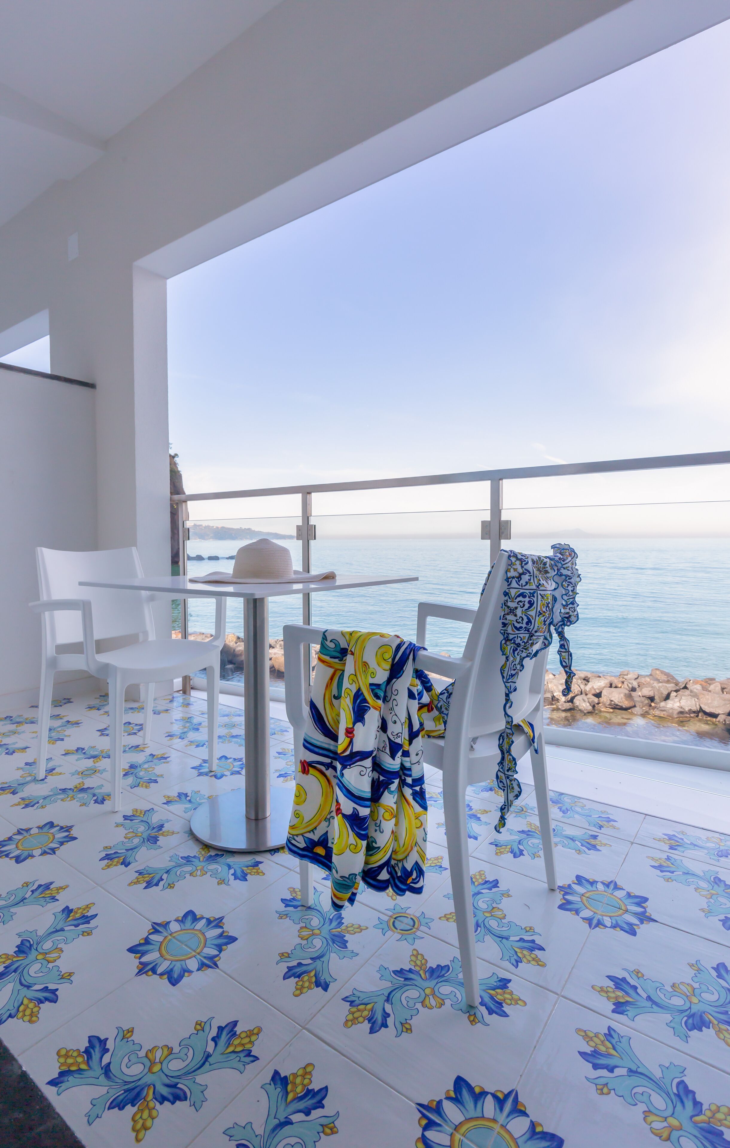 Un balcón con sillas blancas, una mesa y un mantel estampado. El suelo tiene azulejos florales azules y amarillos.